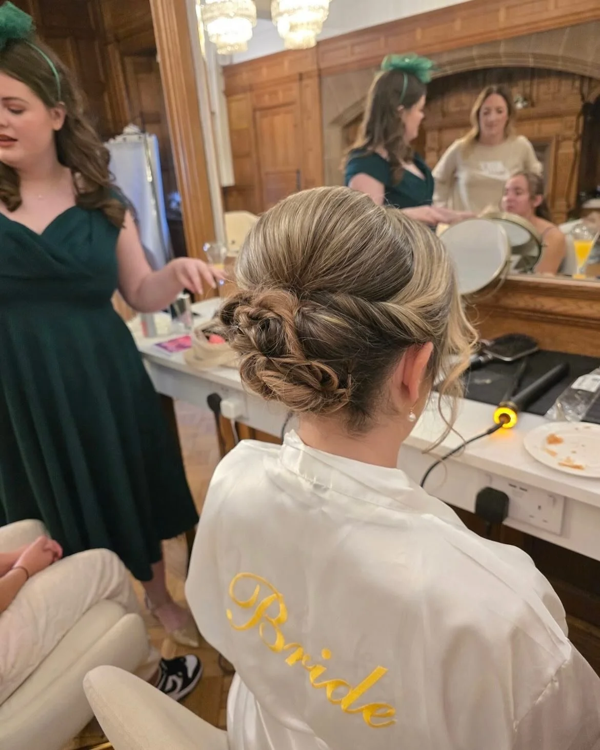 A woman with braided updo hairstyle sitting in front of a mirror, wearing a bridal robe with 'Bride' embroidered at Lisa B Hair & Beauty Salon, Carlisle, Cumbria UK.