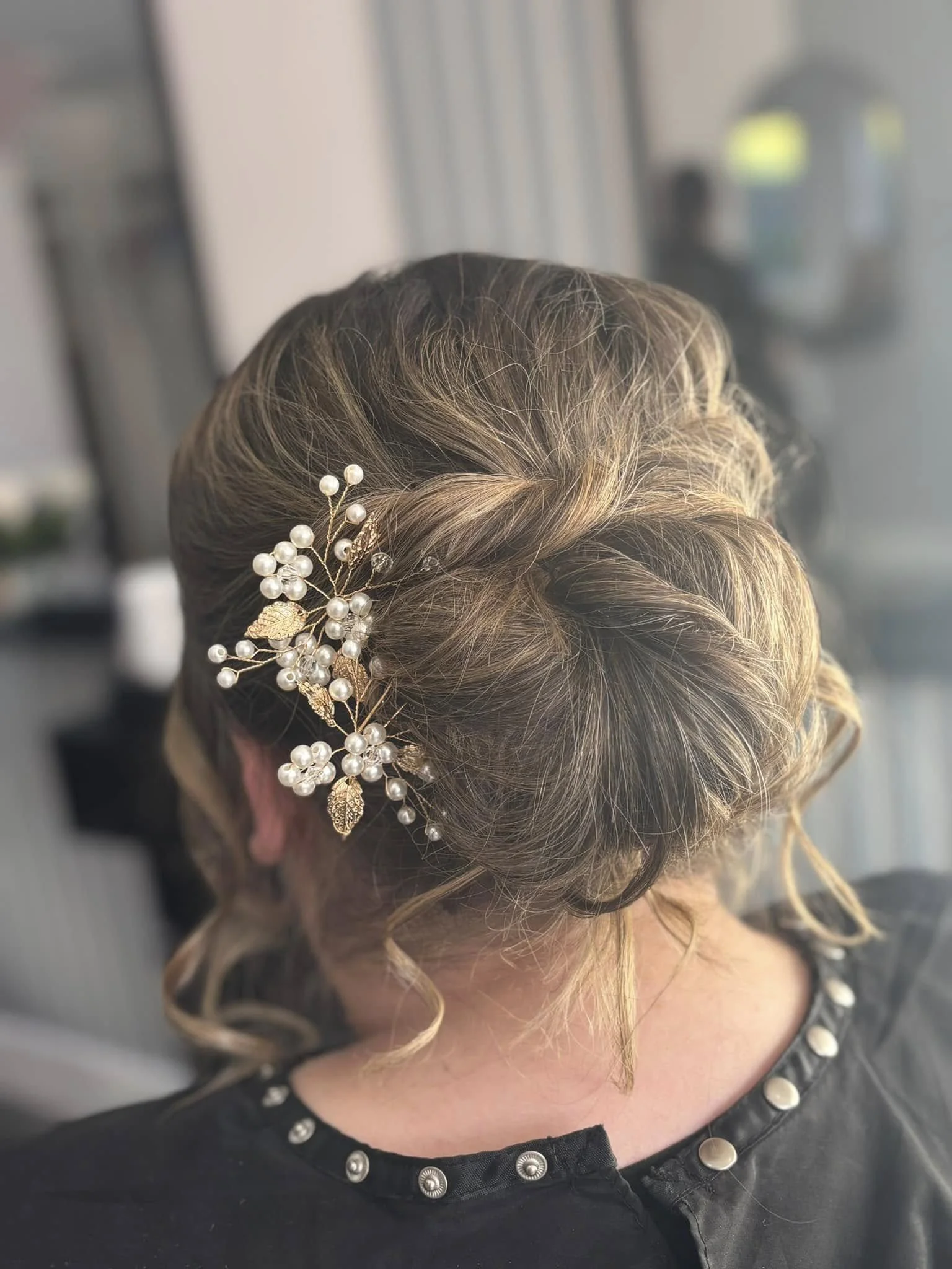 A person's head with styled, blonde and brown hair adorned with a decorative hairpiece featuring pearls and gold leaves, and painted nails with a pearl design. The person is wearing a black top with metal button embellishments.