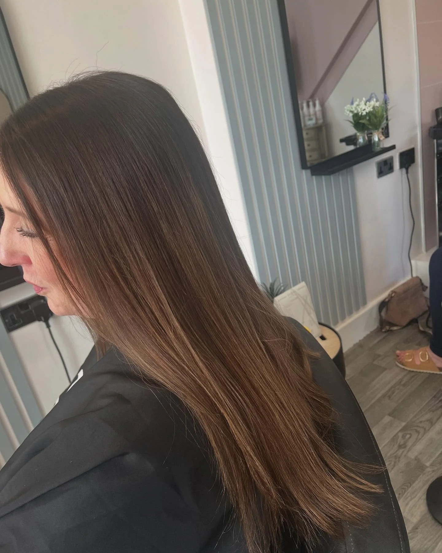 Close-up of a woman with long, straight, brown hair, sitting in a salon chair with a mirror and salon tools in the background.