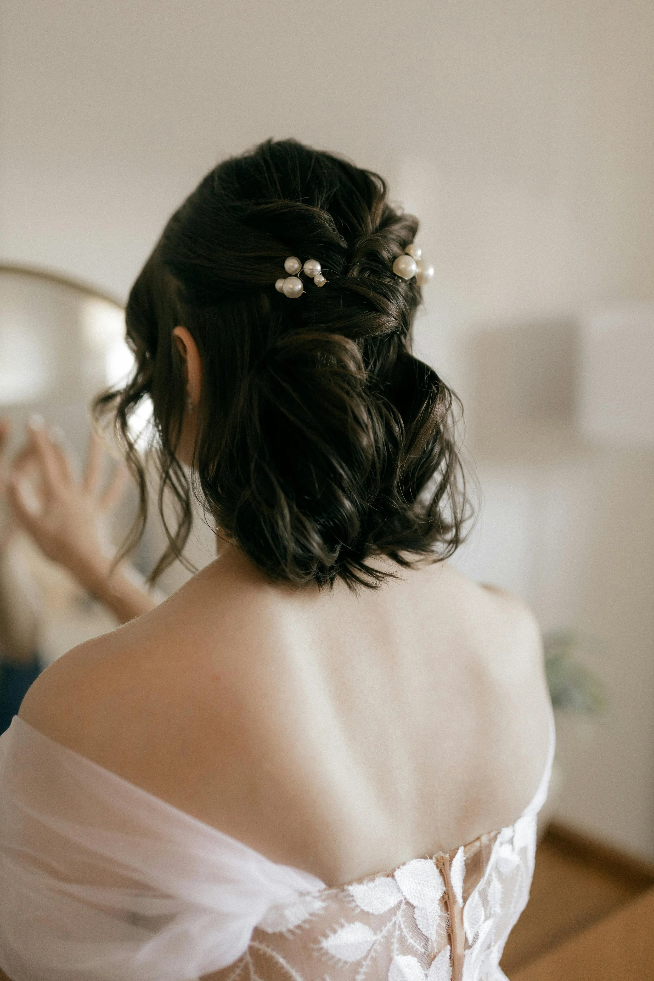 Back view of a woman with dark, curly hair styled with pearl hair accessories, wearing a white off-shoulder lace dress, adjusting her hair.