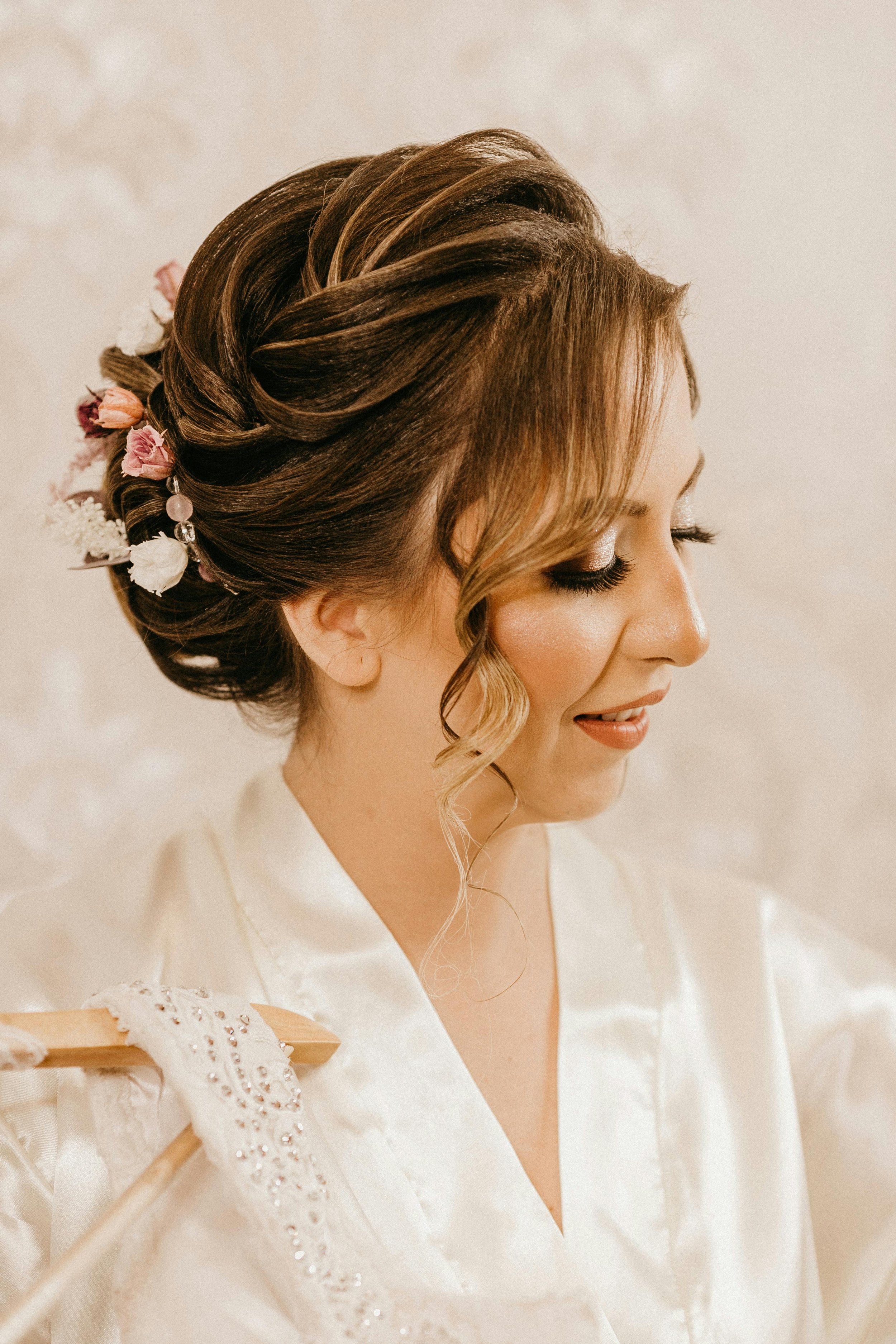 A woman with styled brown hair with highlights, decorated with small pink and white flowers and dangling beads, wearing a white satin robe with lace trim, smiling with her eyes closed, in a softly lit room.
