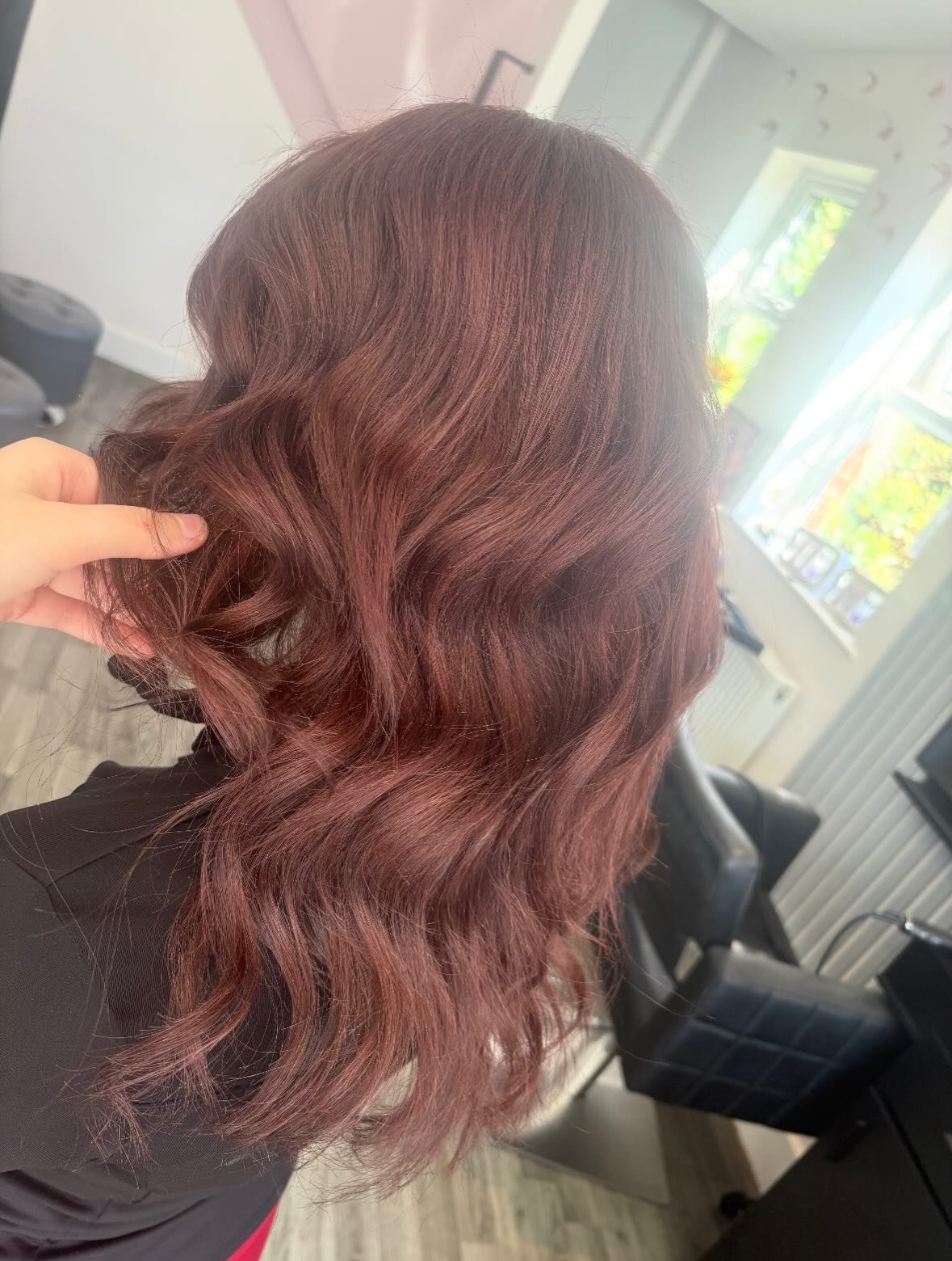 A woman with wavy, reddish-brown hair sitting in a salon chair. A hand is holding some of her hair, showing its texture and color. Natural light comes from a window in the background.