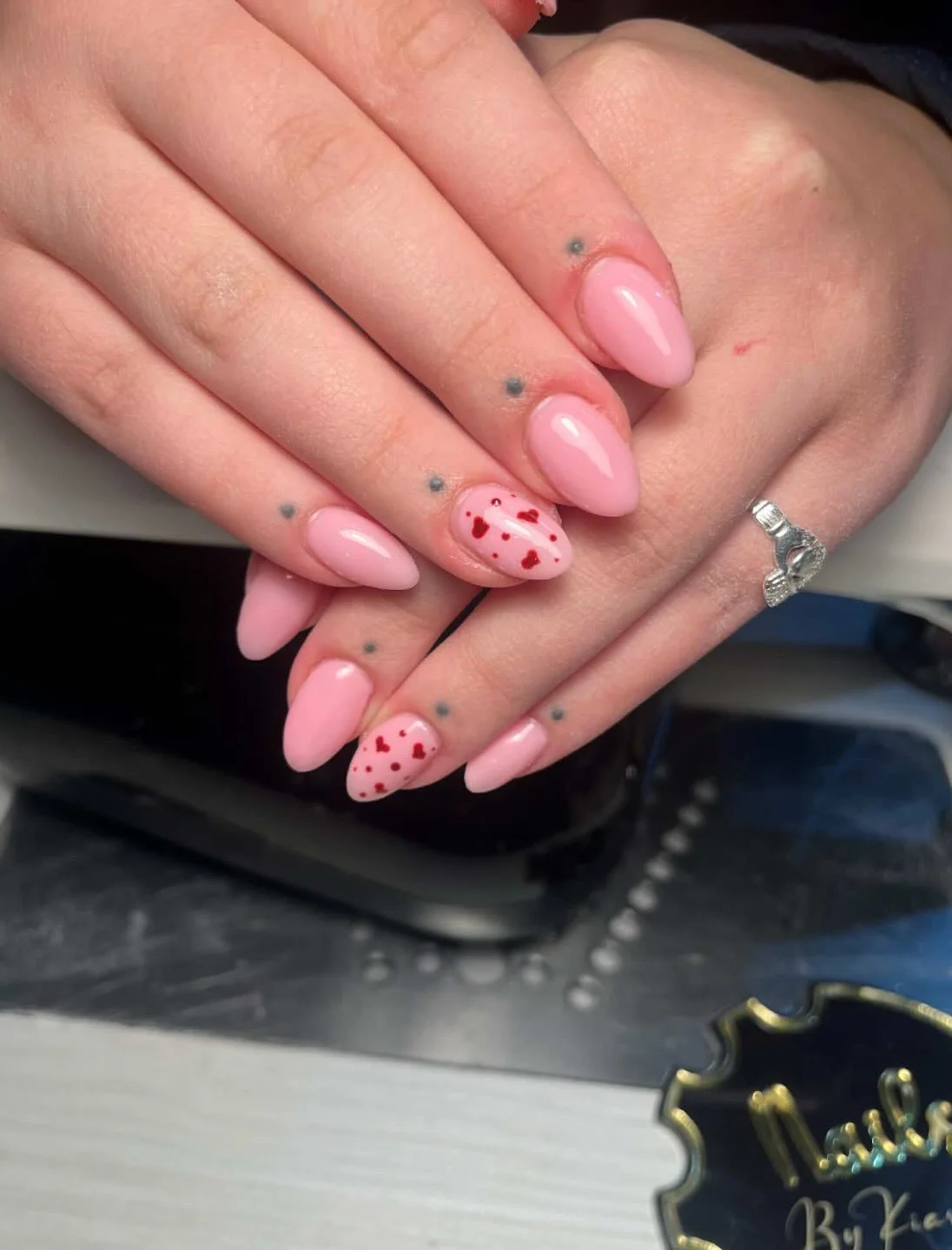 Close-up of manicured nails with light pink polish and decorative hearts on some nails, with a silver ring on the finger, resting on a black and white surface.