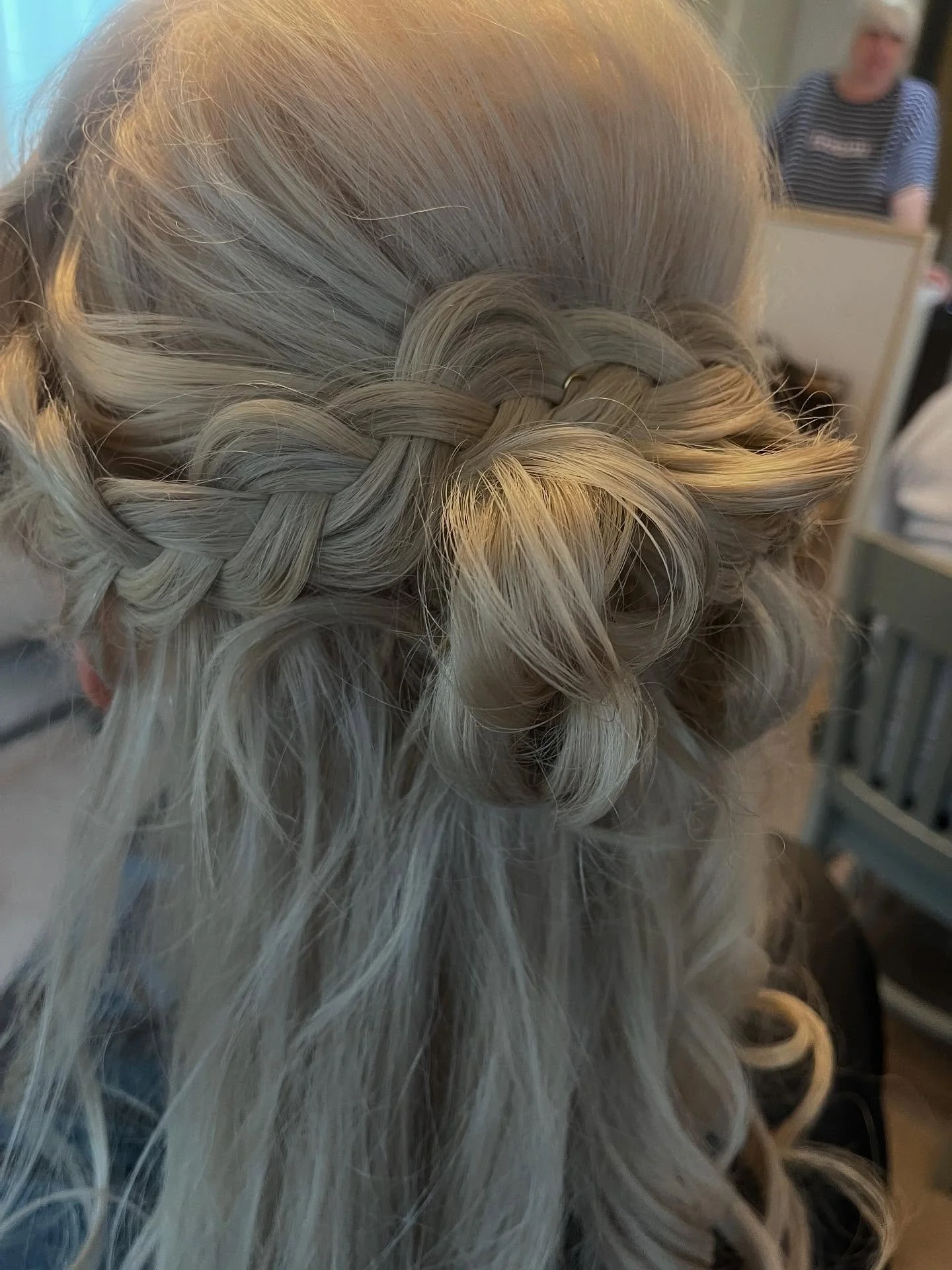 Close-up of a person's hair styled in a crown braid with loose curls, with a woman wearing a striped shirt in the background.