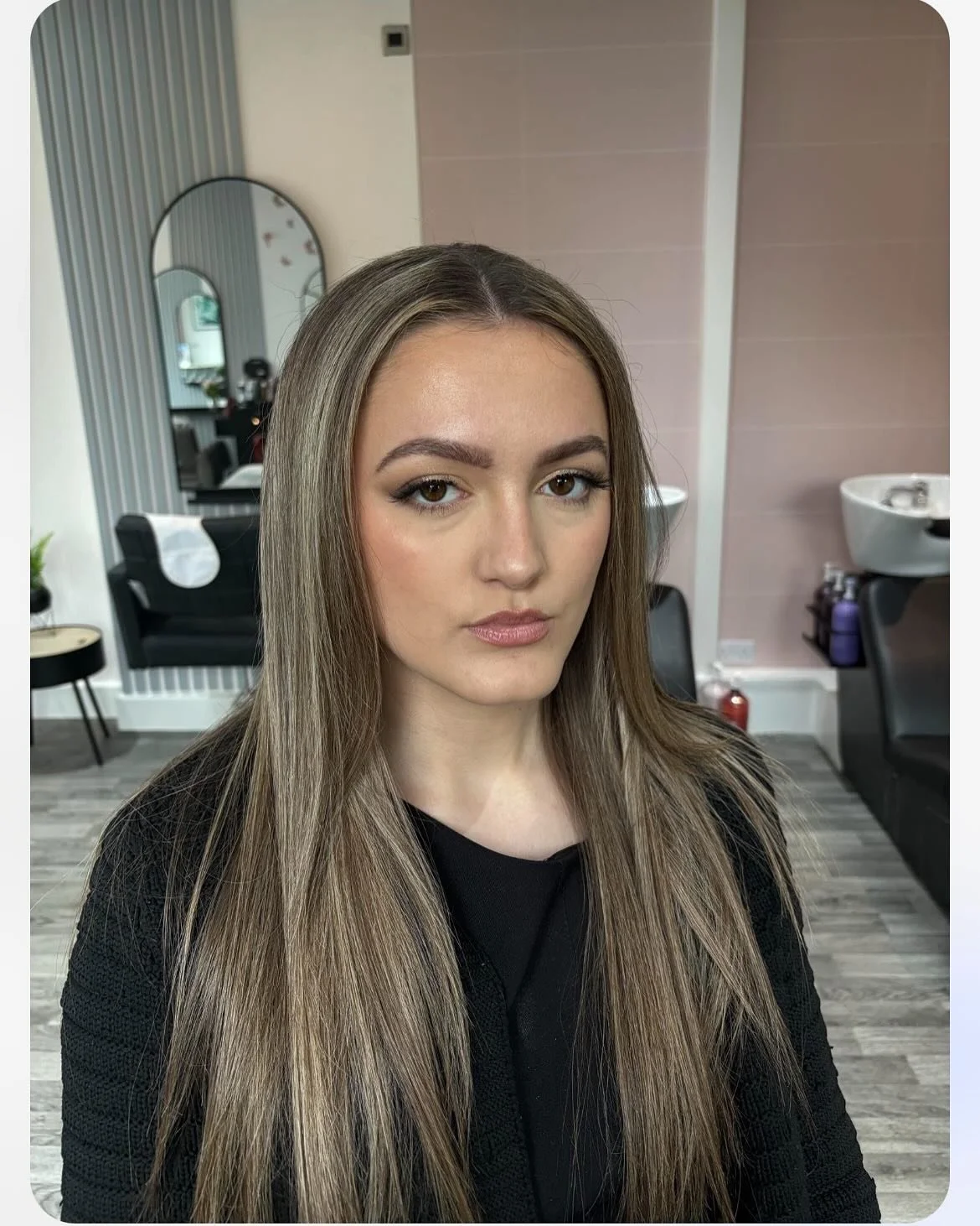 A young woman with long, straight, light brown hair looking at the camera, inside a salon with mirrors, chairs, and hair washing basins in the background.