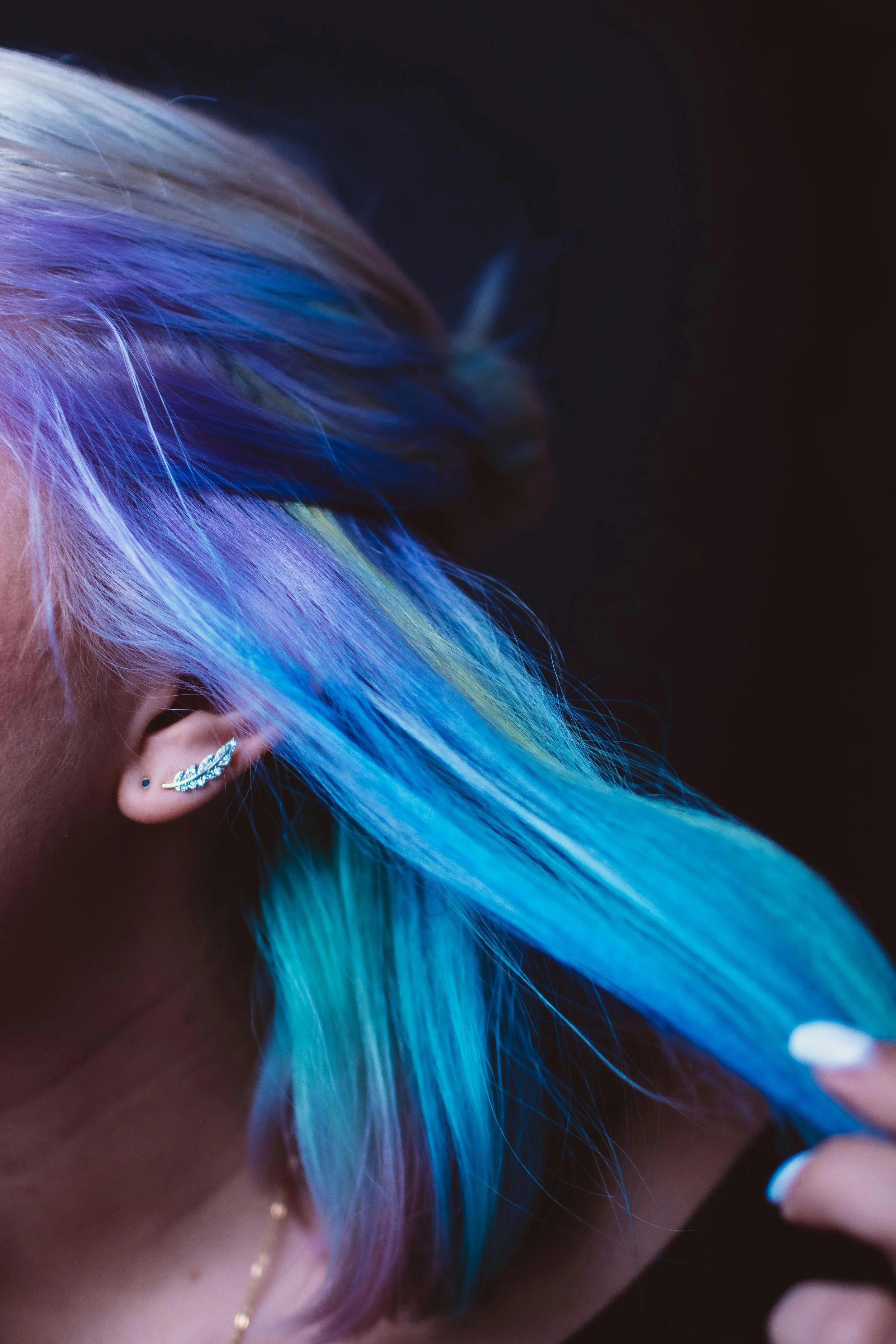 Close-up of a person's ear with a feather earring and vibrant blue and purple hair against a dark background.