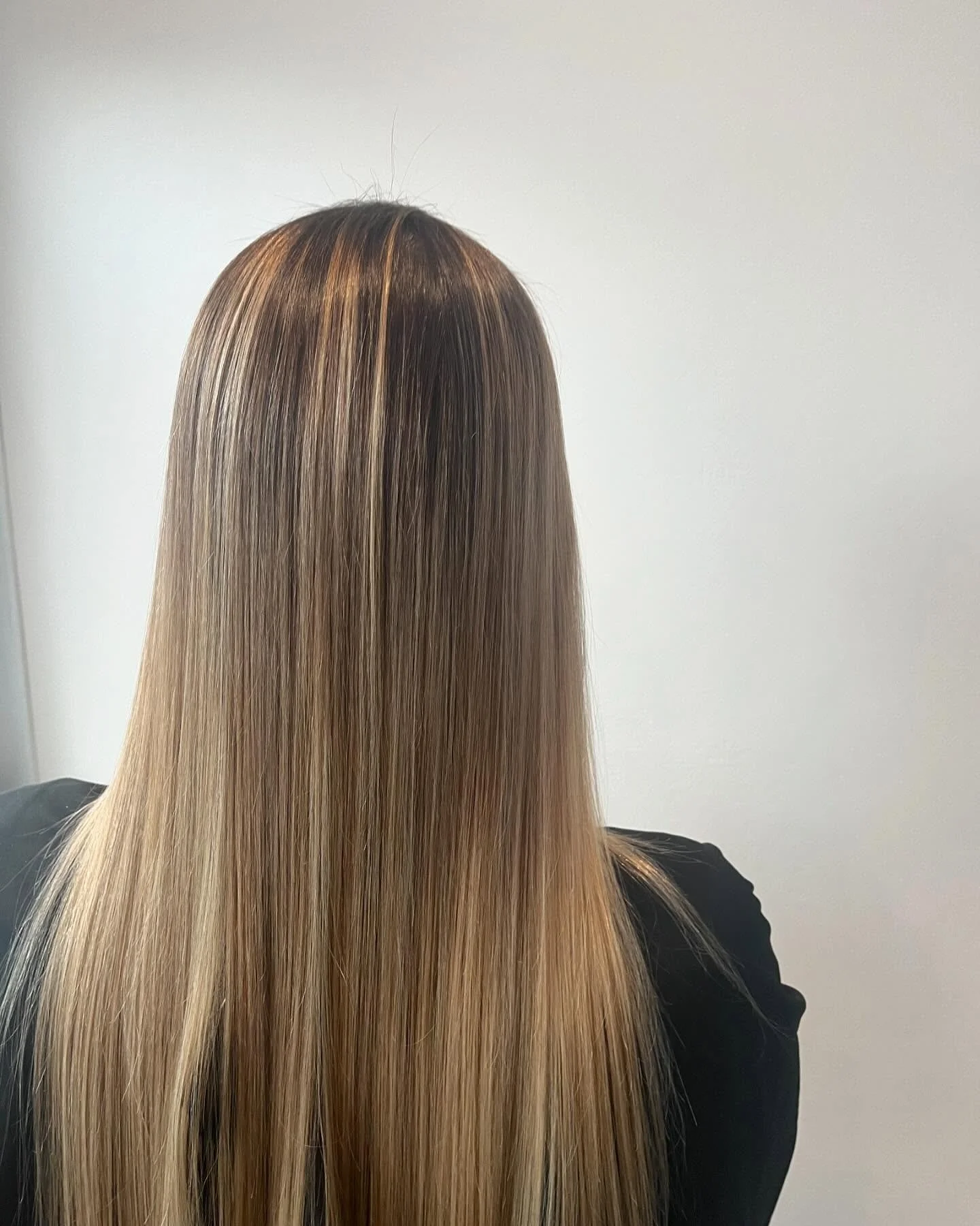 Back view of a person with long, straight blonde hair, wearing a black top, against a plain white wall.
