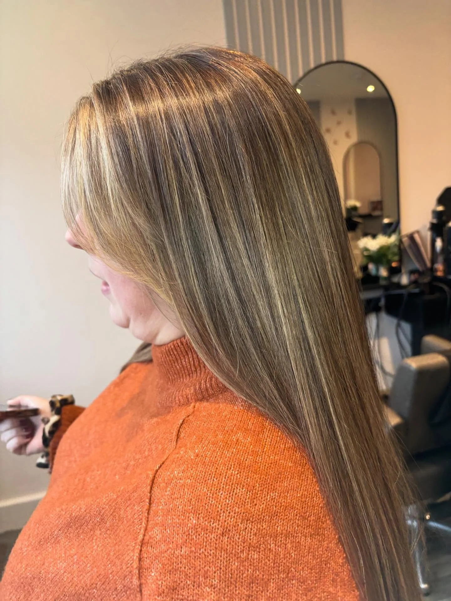 A woman with long, straight, shiny blonde hair, wearing an orange sweater, is in a salon or similar setting. Lisa B Hair & Beauty Salon, Carlisle, Cumbria UK.