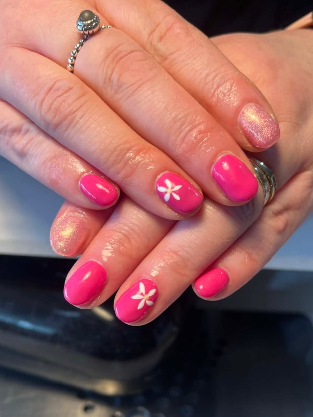 Close-up of a person's hands with pink and glittery nail polish, some nails featuring white flower designs, and a silver ring on one finger.