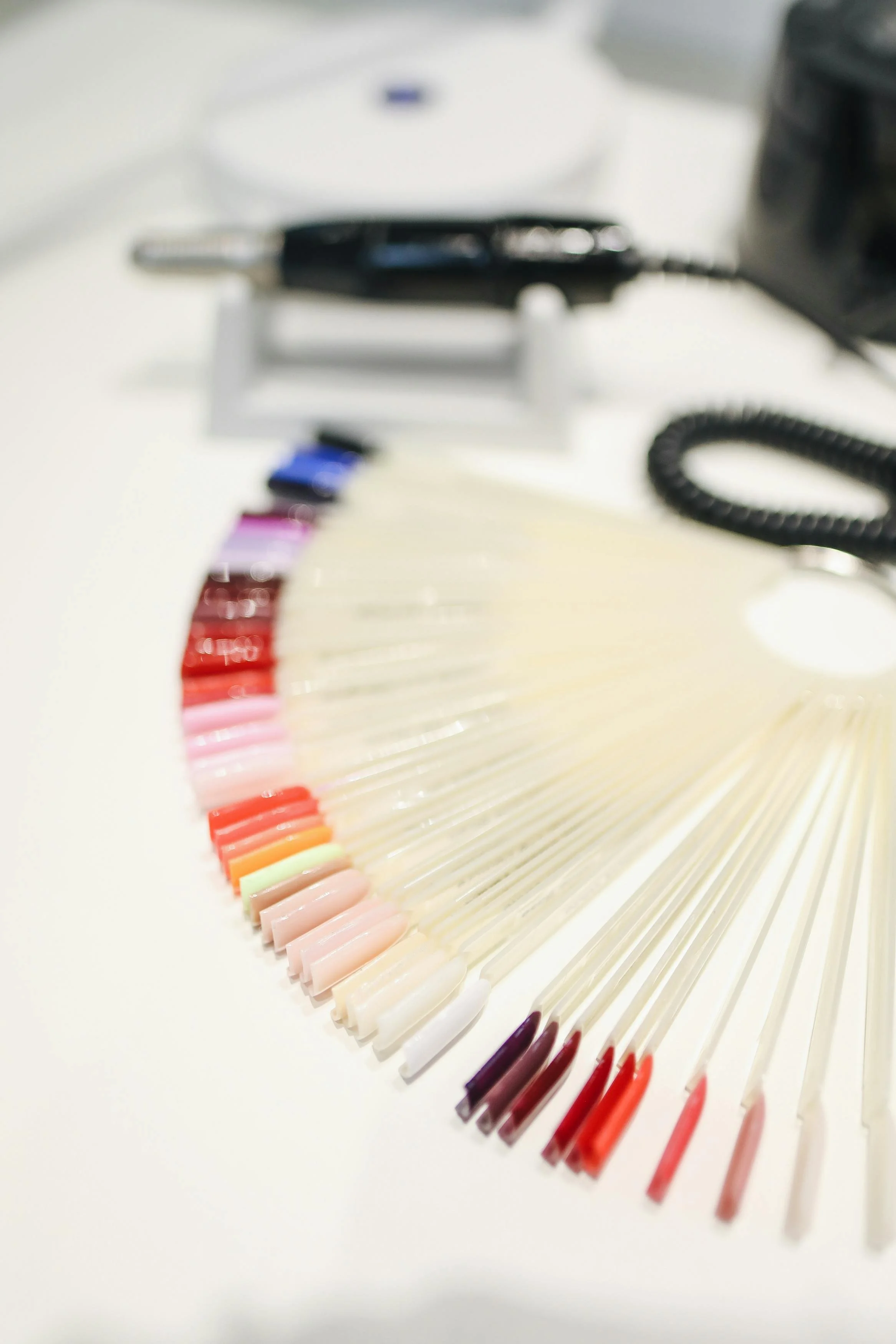 Color swatches of nail polish on sample sticks arranged in a semi-circle, with a display of nail polish colors and a black cord in the background.