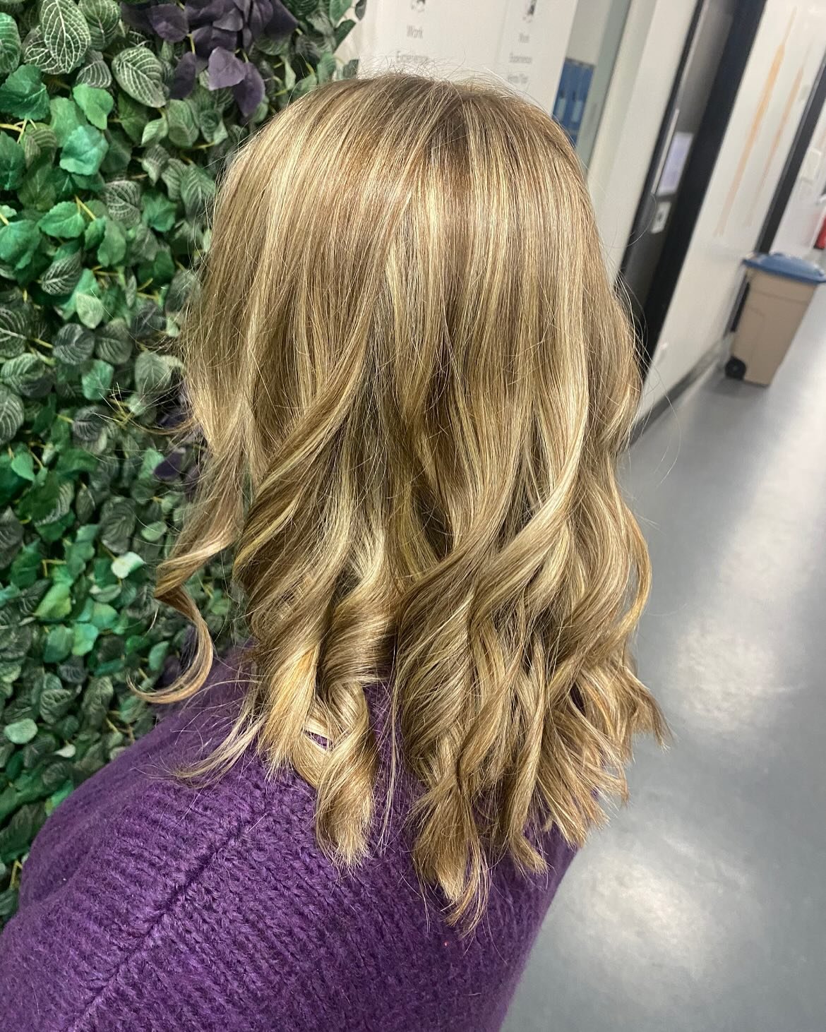 Back view of a person with shoulder-length, wavy blonde hair, wearing a purple sweater, standing near a wall with green foliage.