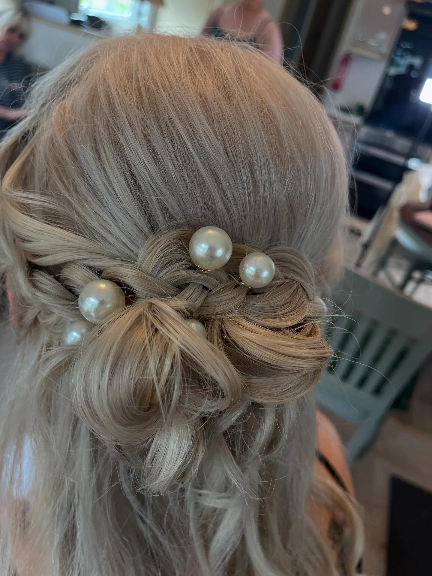 Blonde hair styled in an elegant updo decorated with large pearl pins, in a setting that appears to be a hair salon with blurred people and equipment in the background.