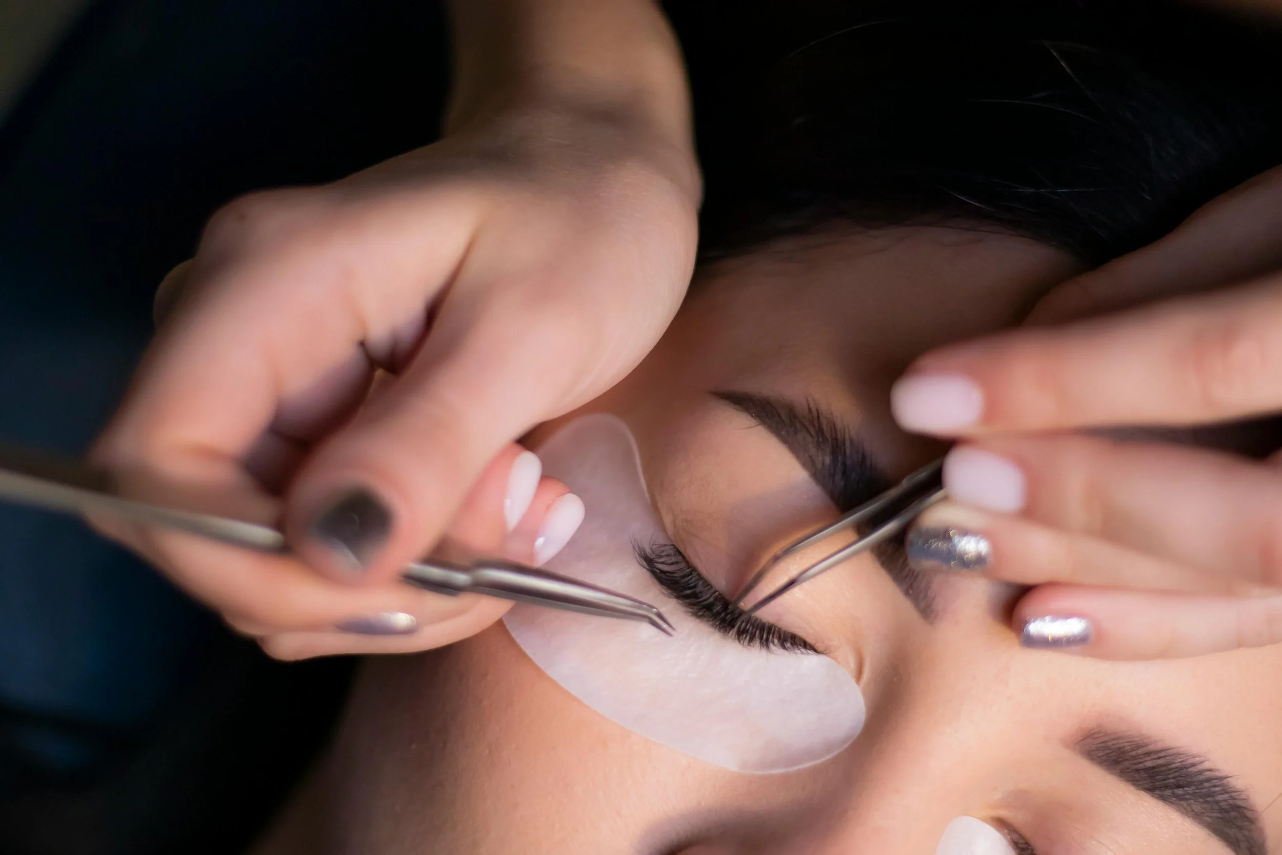 Close-up of a person receiving eyelash extensions from a technician using tweezers, with the person's eye closed.