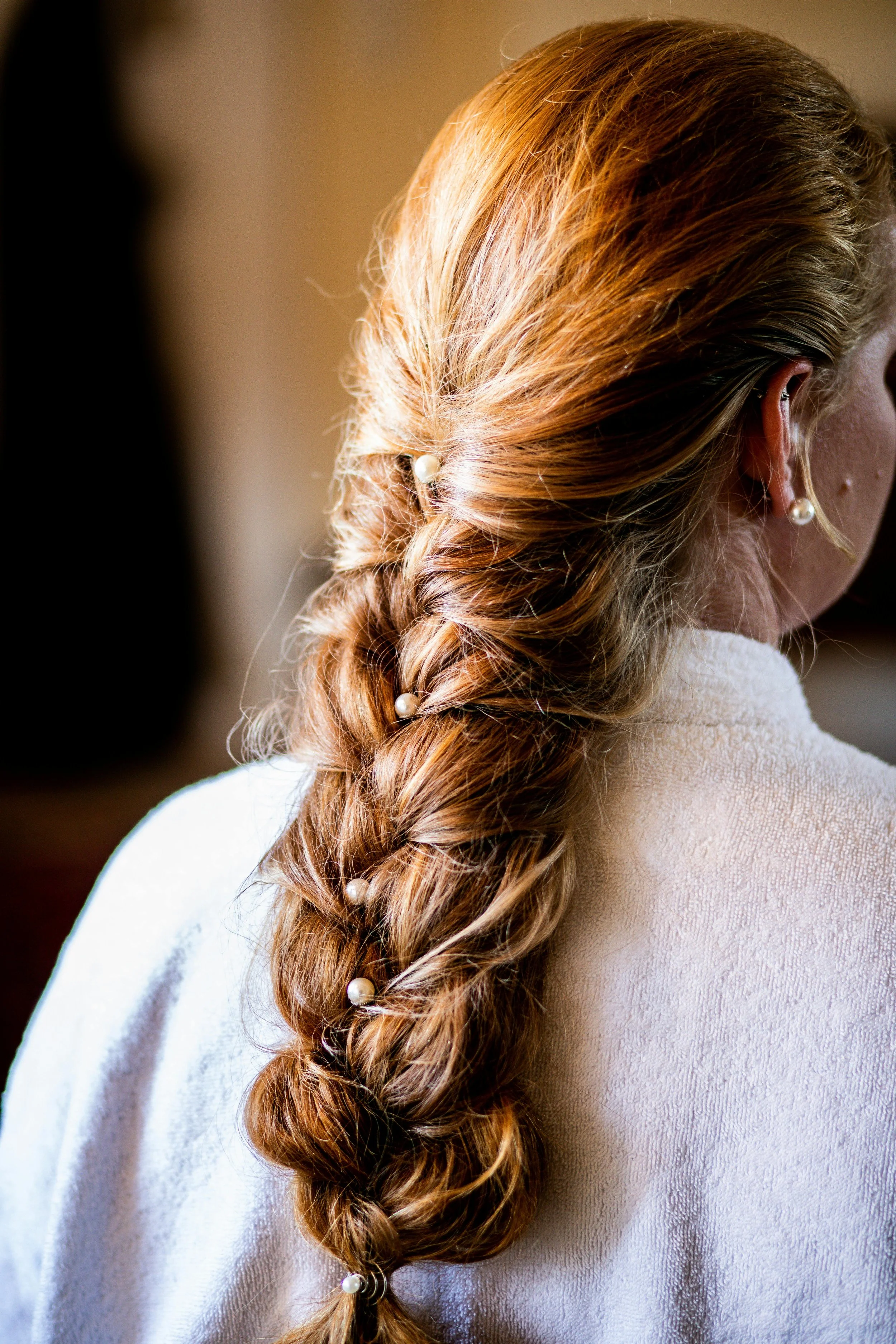 Close-up of a woman with red, wavy hair styled in a loose braid, adorned with pearl hairpins, wearing pearl earrings, and dressed in a white textured garment.