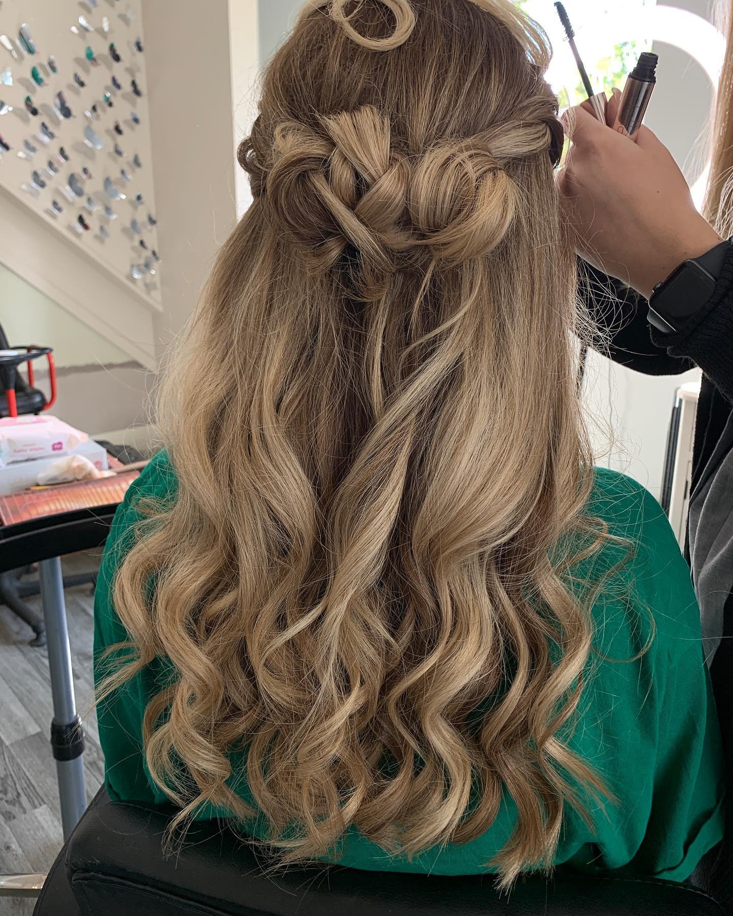 Back view of a woman with long blonde wavy hair, styled with a braided crown, sitting in a salon chair, getting her hair done by a stylist.