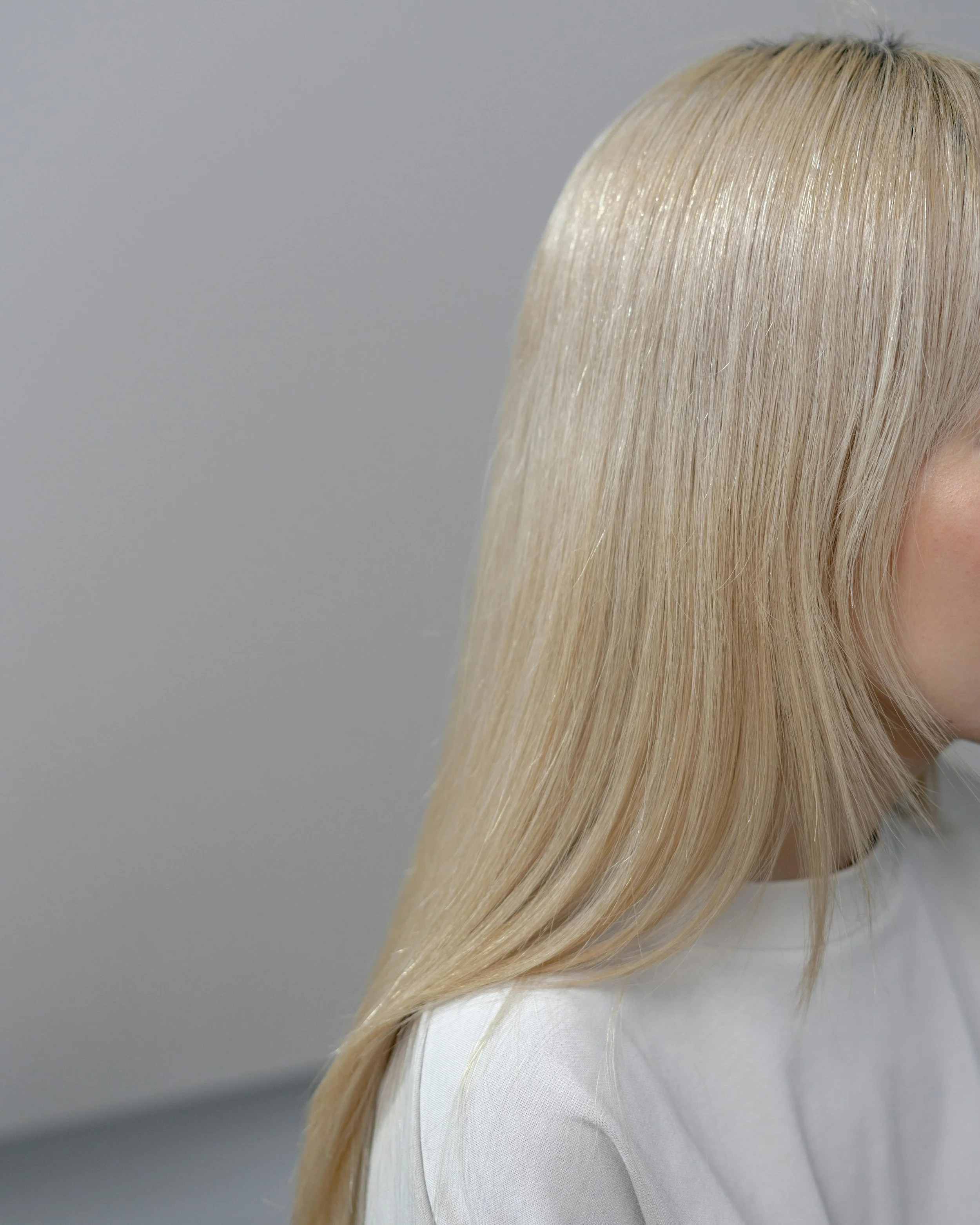 Close-up of a blonde woman's smooth and shiny hair, partially covering her face, against a plain gray background.