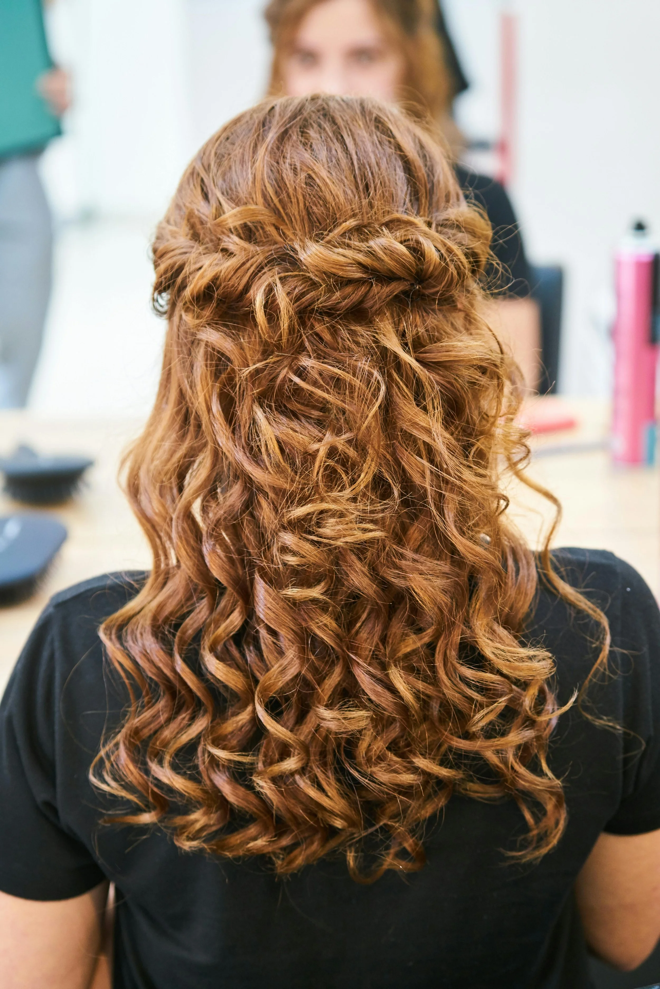 Close-up of a woman with long, curly, reddish-brown hair in a half-up hairstyle, sitting in front of a mirror, wearing a black top, in a hair salon.
