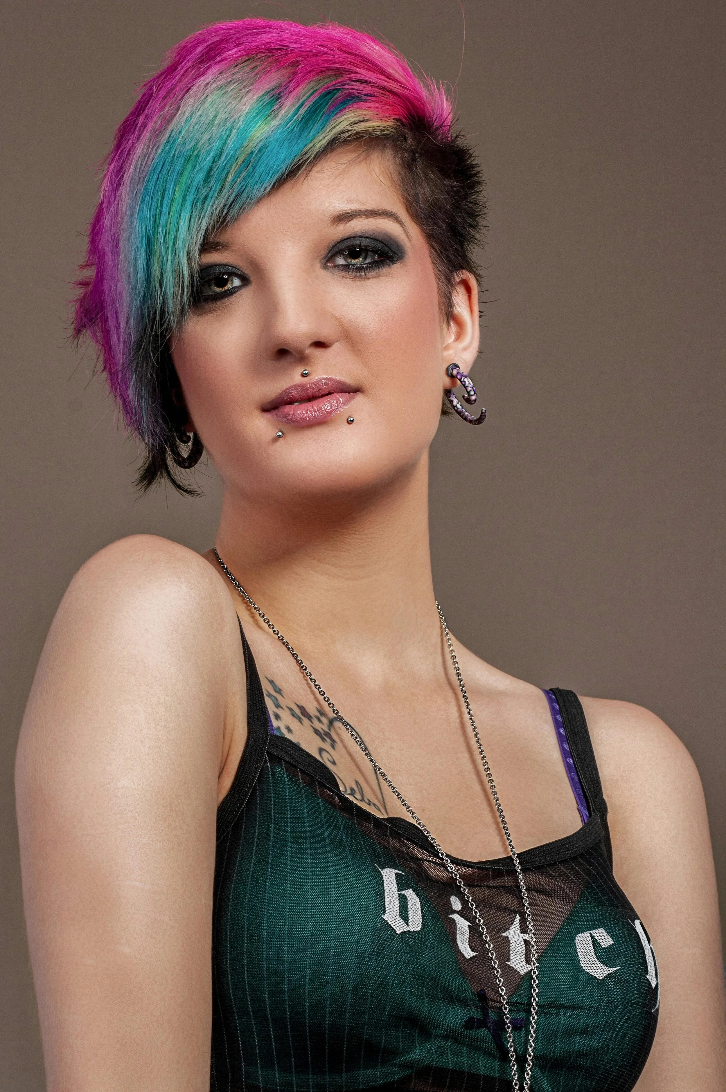 A woman with short, multicolored hair including pink, blue, green, and gray shades, wearing dark makeup and jewelry, posing against a plain background.