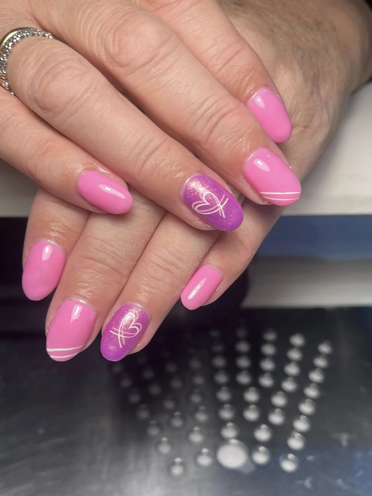 Close-up of hands with pink manicured nails, some with white stripe designs and heart accents, resting on a perforated metallic surface. One hand has a silver ring.