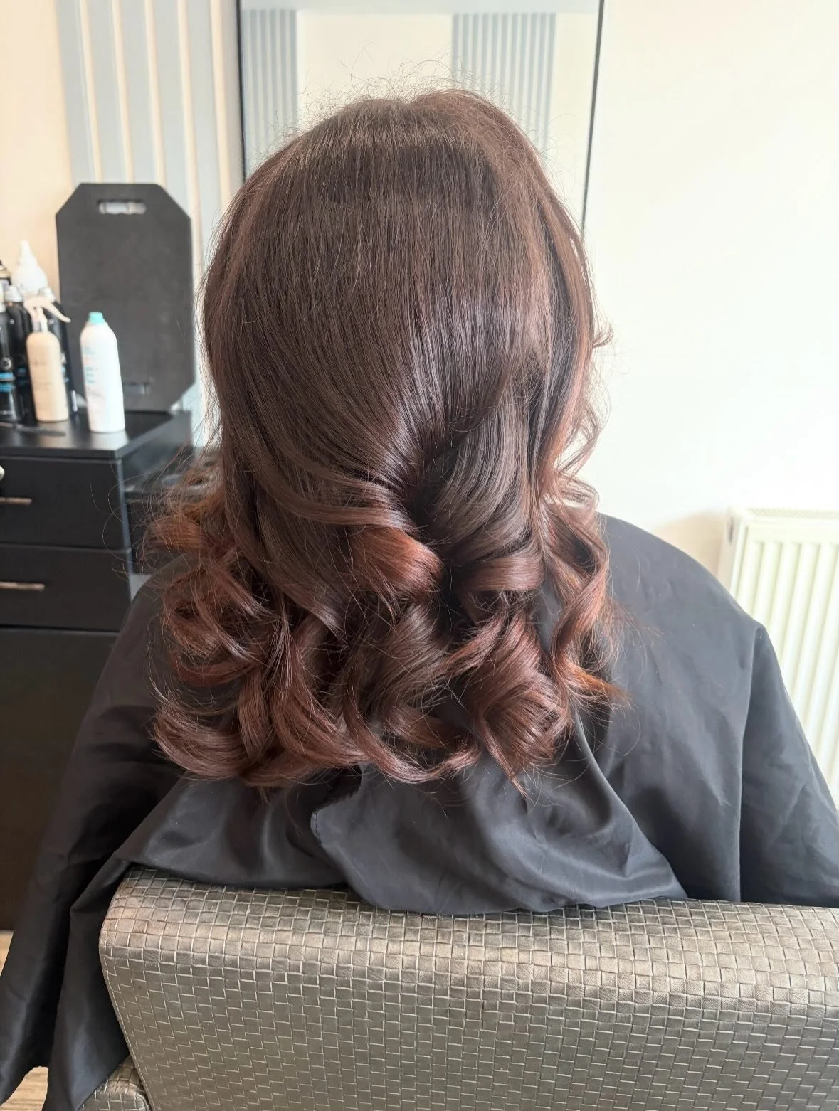Woman with medium-length hair styled in loose curls, sitting in a salon chair. The hair is a rich brown with subtle highlights. Salon bottles and tools are visible in the background.