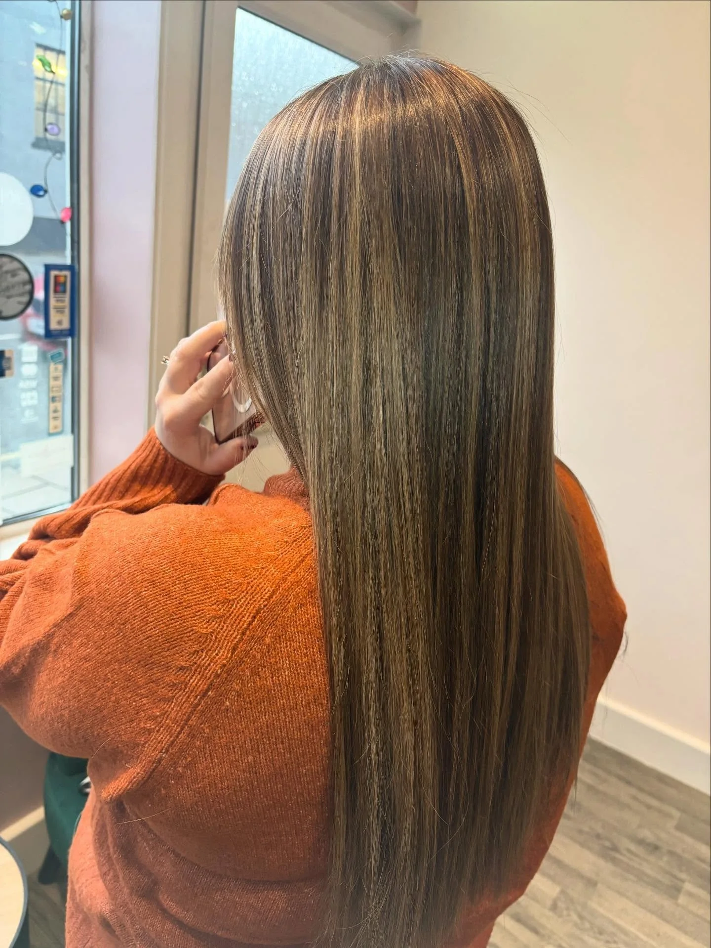 Back of a woman with long, straight, blonde hair in a salon, wearing an orange sweater, near a window with a blurred background.
