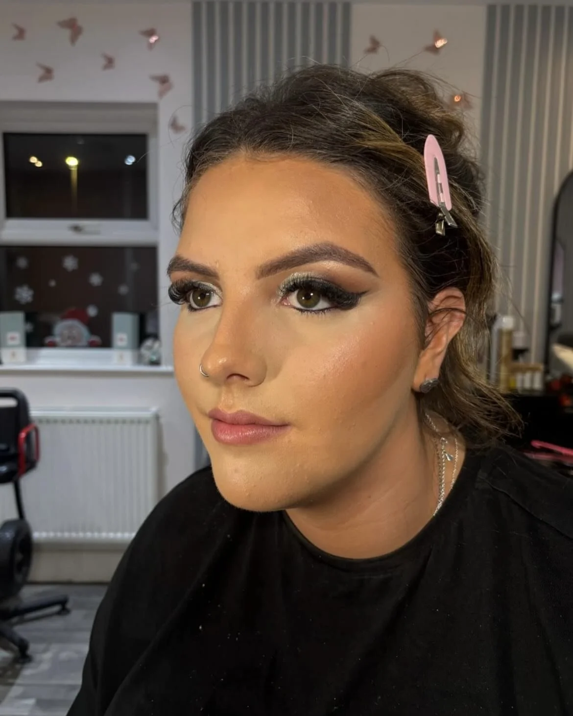 A woman with makeup and a nose piercing, sitting indoors with Christmas decorations visible in the background, including snowflake decals and a Santa figure on the window.