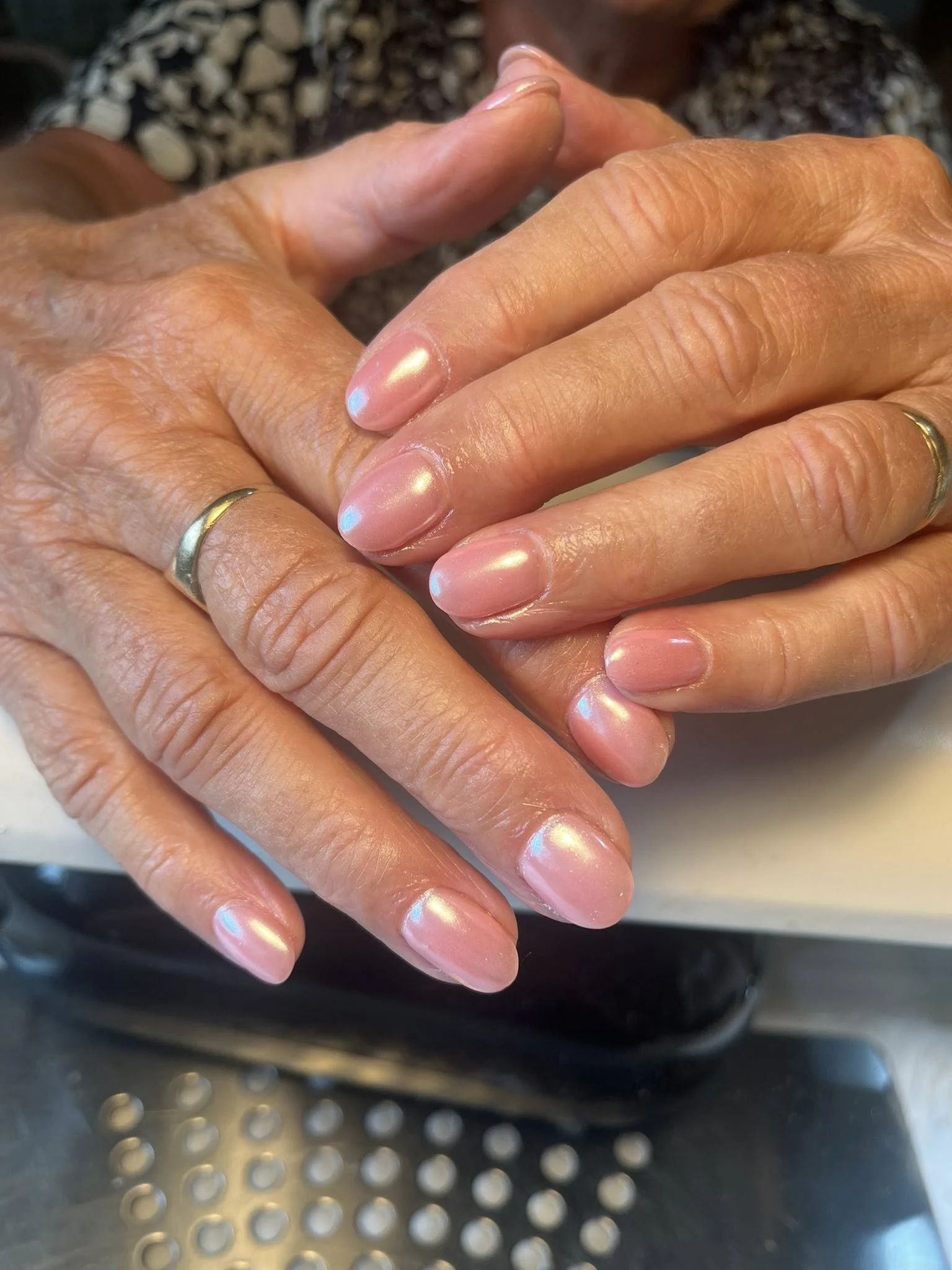 Close-up of two hands clasped together, with shiny, pearlescent nails and rings on the fingers, resting on a perforated metal surface.