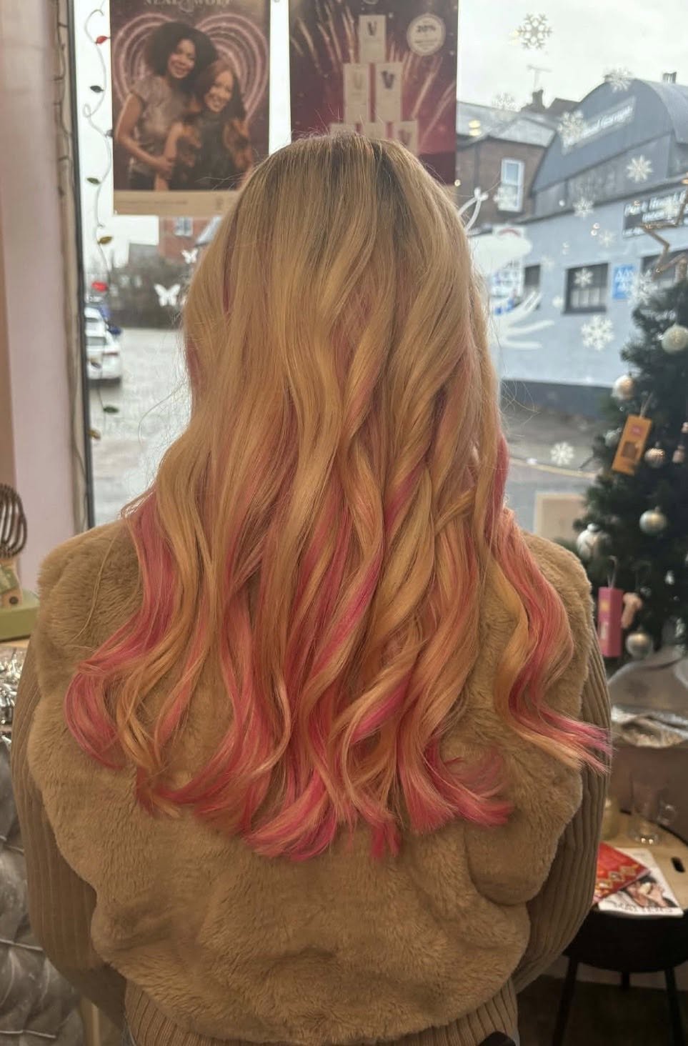Back view of a woman with long wavy hair dyed pink and blonde, standing inside near a window with snowflake decorations and a Christmas tree, with posters and buildings visible outside.