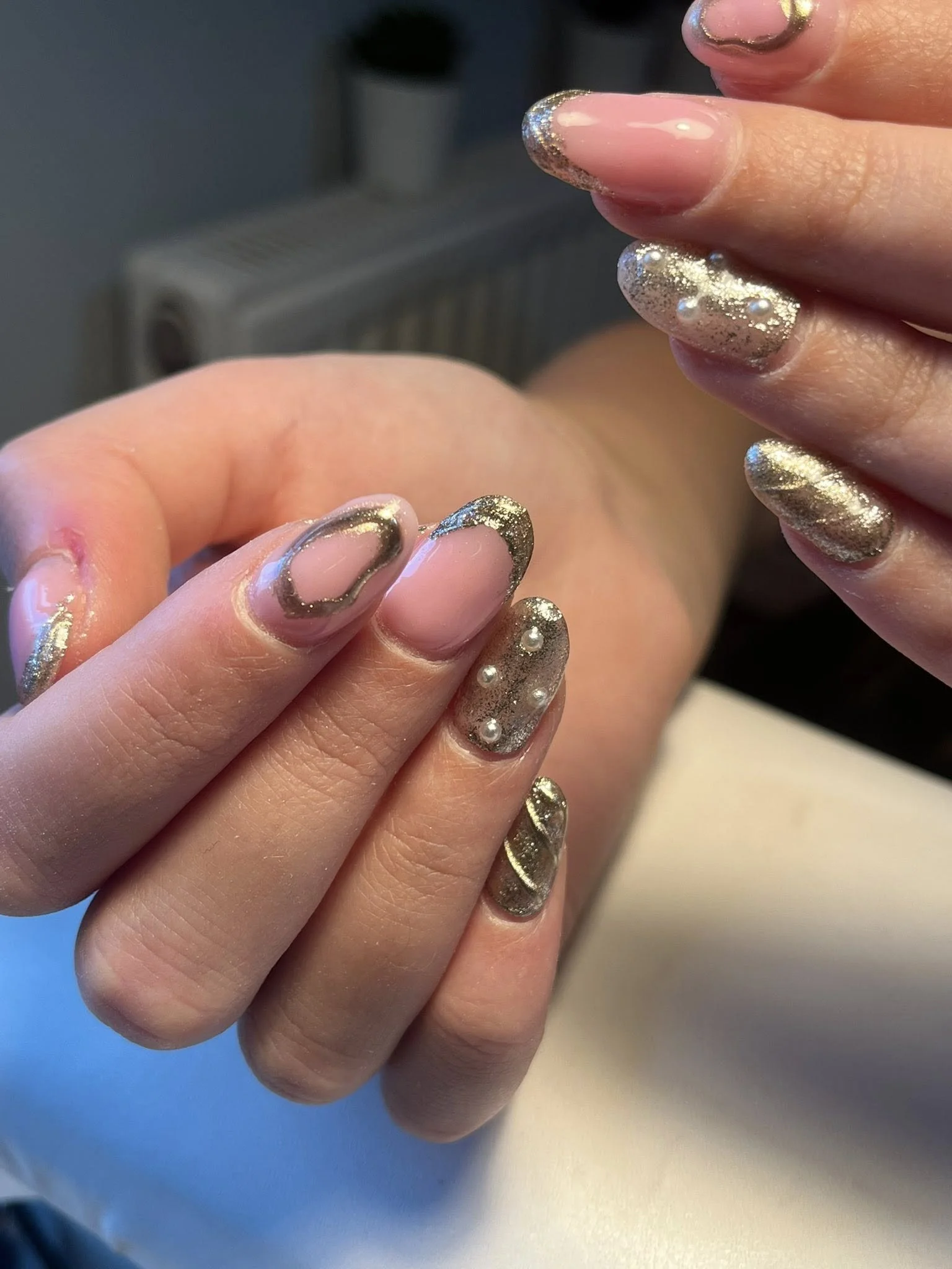 Close-up of a person's hand with nails painted in a nude base color and decorated with gold glitter and small white pearls.