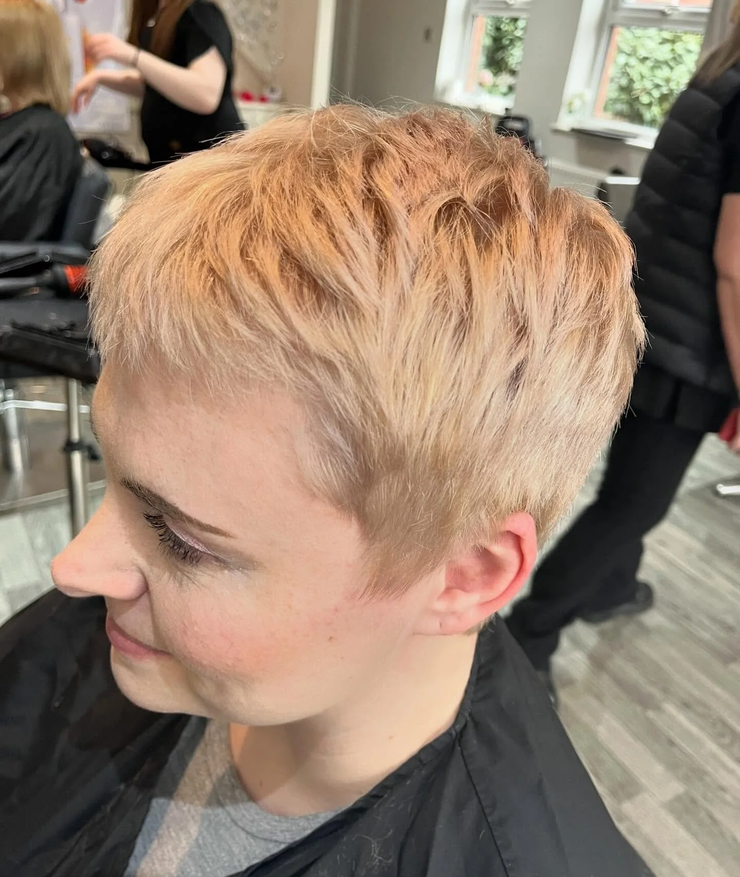 A person with short, blonde, reddish hair in a salon. In the background, a hairstylist and another customer are visible.