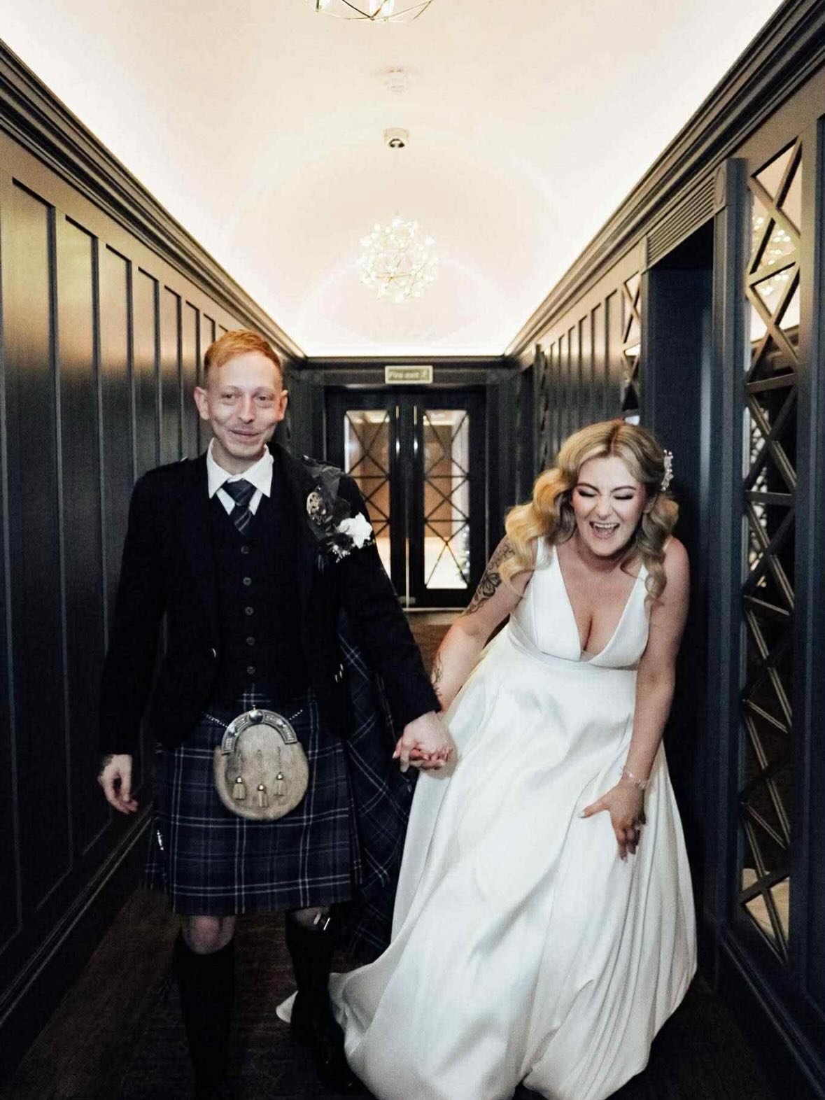 A happy couple in wedding attire holding hands and walking down a hallway, with the bride laughing and the groom smiling. Lisa B Hair & Beauty Salon, Carlisle, Cumbria UK.