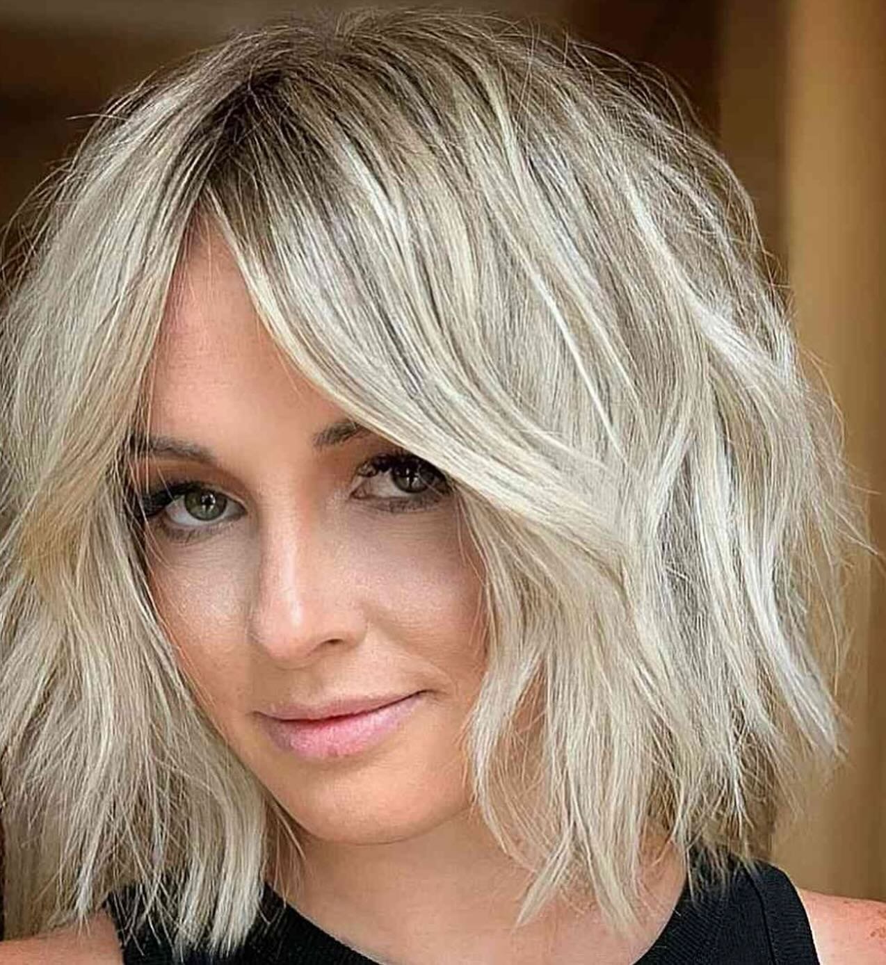 Close-up of a woman with shoulder-length, layered, blonde hair and green eyes, wearing makeup and a black top.