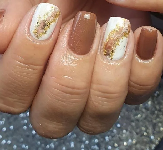 Close-up of a hand with manicured nails painted in white and brown, featuring gold foil accents. Award winning Lisa B Hair and Beauty Salon in Carlisle, Cumbria UK.