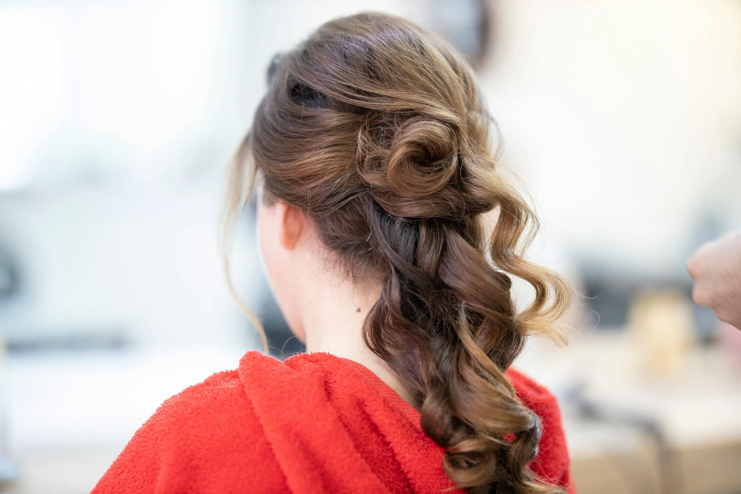 A woman with brown hair styled in loose curls, wearing a red garment, is seen from behind with a soft background.