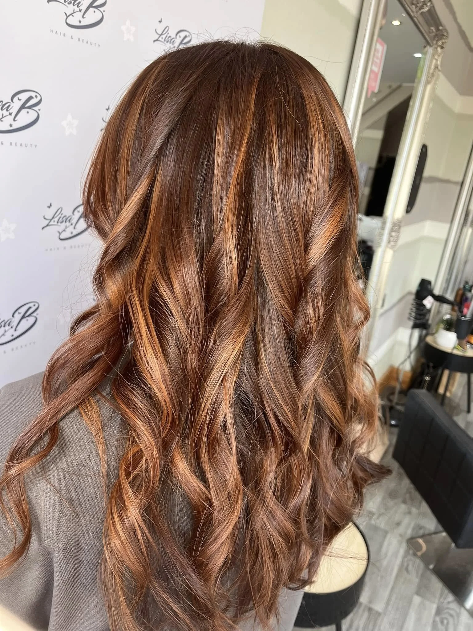 A person with long, wavy, reddish-brown hair with highlights, sitting in a salon with a mirror and salon equipment in the background.