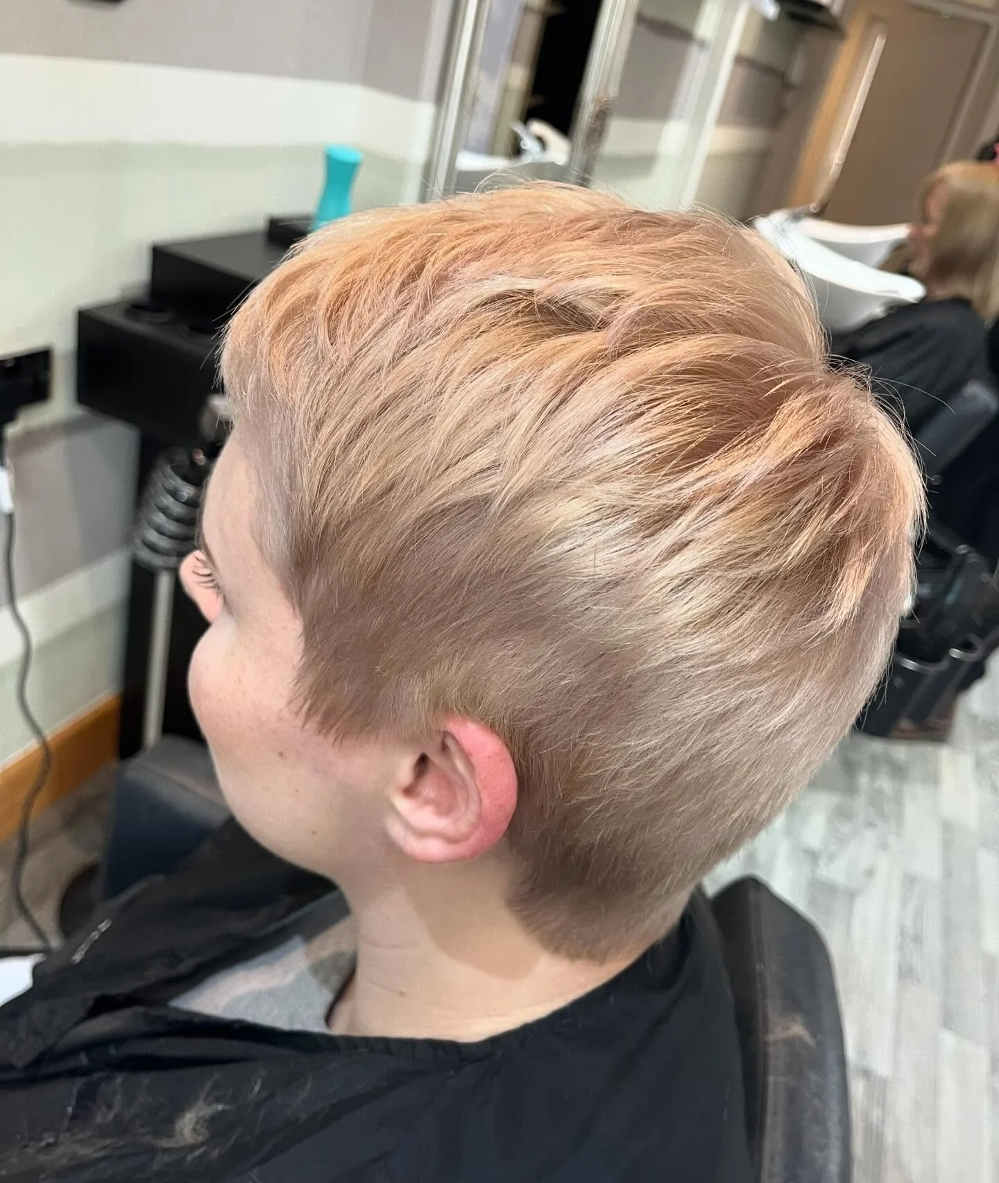 Young person with short, blonde hair sitting in a hair salon chair, receiving a haircut or styling from a hairstylist.