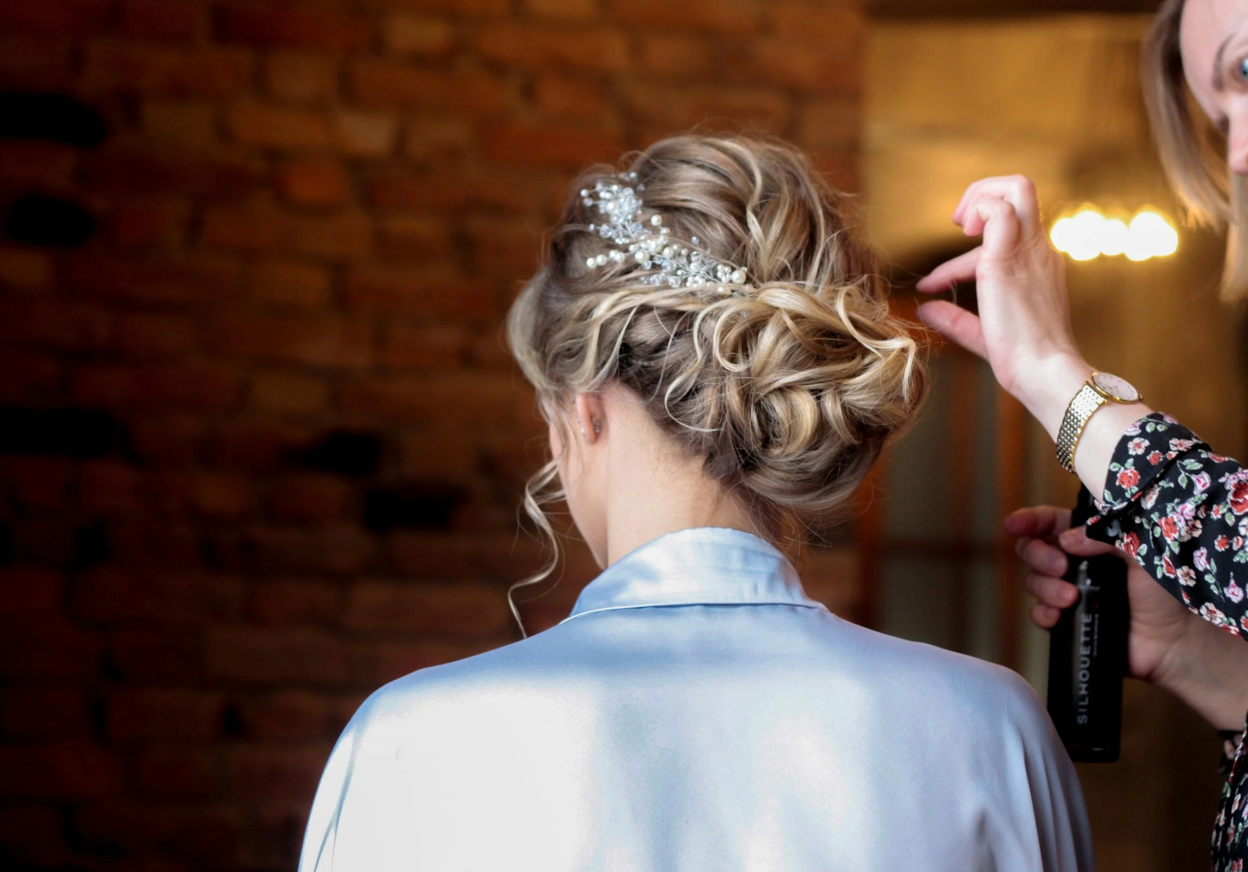 Close-up of a woman with blonde curly hair, adorned with a decorative hair accessory, getting her hair styled by a person wearing a watch and floral shirt, against a brick wall background.