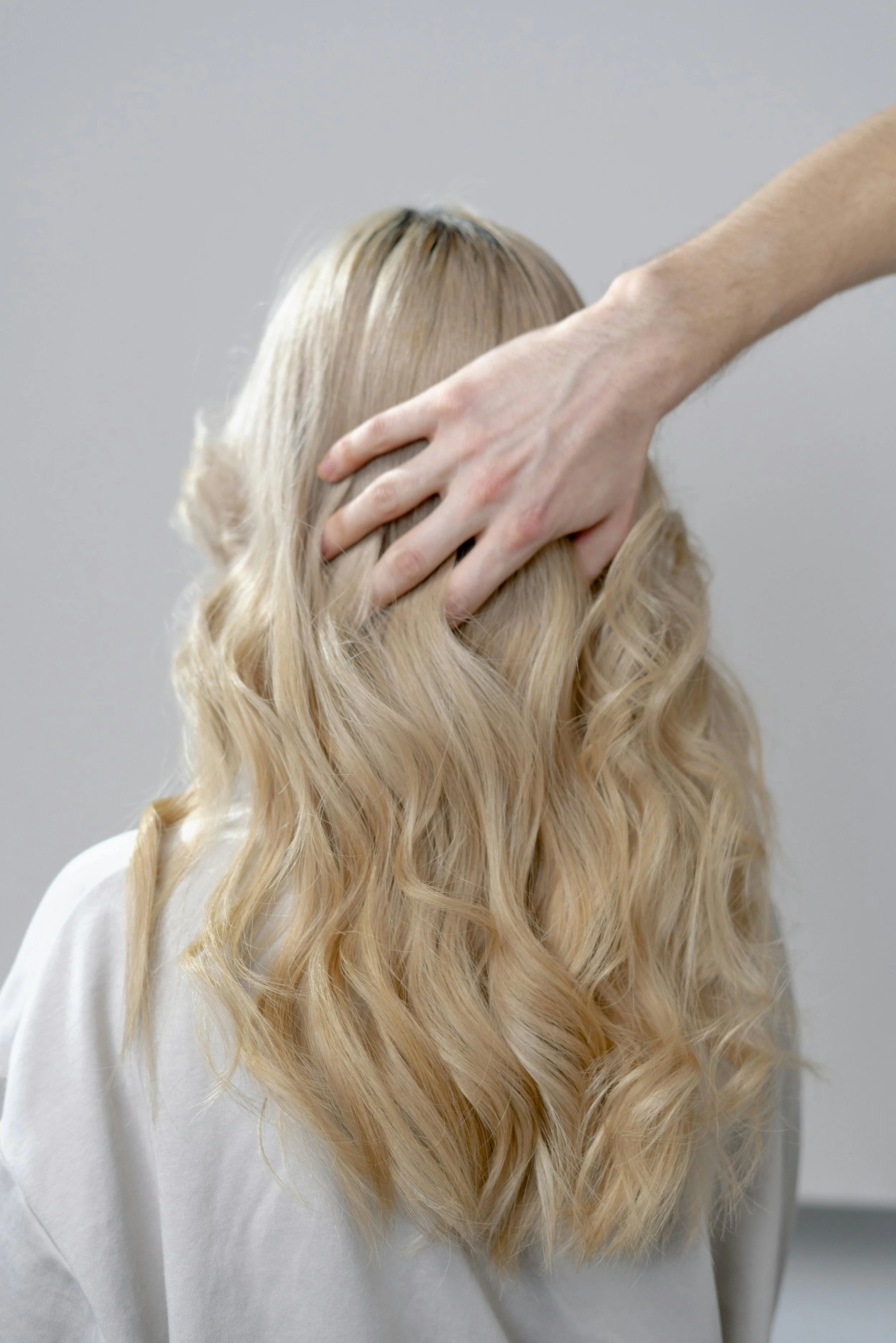 A person with long, wavy blonde hair wearing a white top, touching their scalp with their hand, standing against a plain gray background.