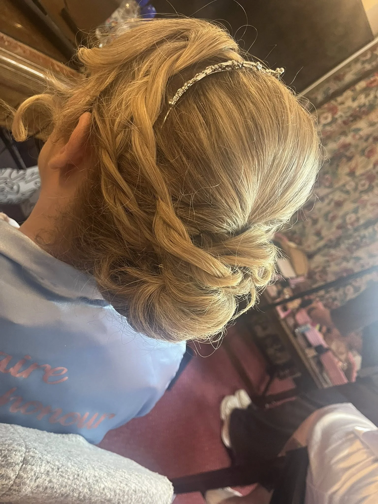 A woman with blonde hair styled in curls wearing a silver tiara, sitting in a room with a floral carpet and a dark wooden dresser in the background.