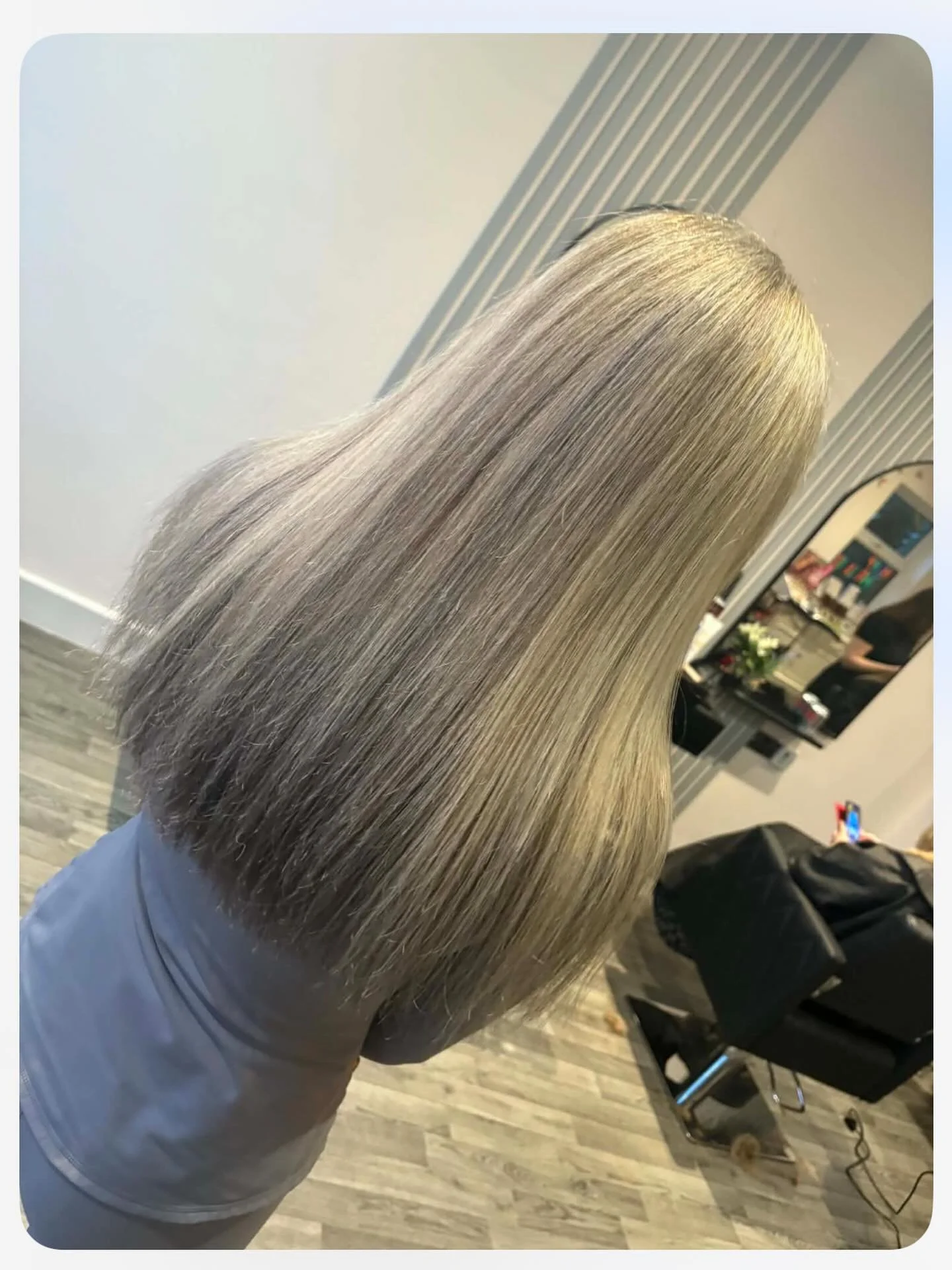 A woman with long, straight, blended gray and blonde hair is sitting in a salon chair, with hair styled smoothly and shiny. In the background, a mirror and salon tools are visible.