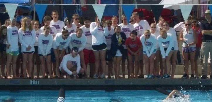 Four young female swimmers wearing swimming caps and goggles standing outdoors at a swimming event, holding a banner that reads '2024-2025 CHAMPION FIRST'. They are smiling and standing on a deck with a pool or water area to their left, with other people gathered in the background.