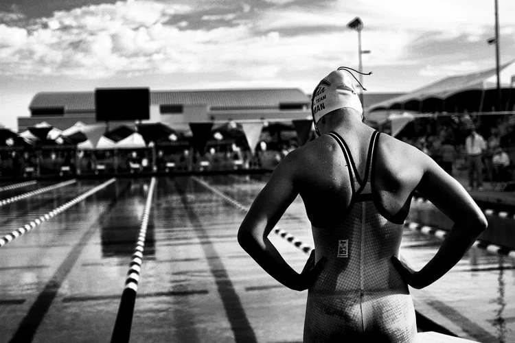 Four young female swimmers in swim caps and goggles smiling together at an outdoor swimming pool.
