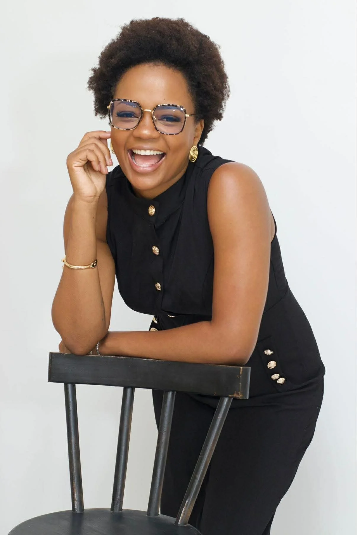 A woman with short curly hair wearing glasses and a black sleeveless dress, smiling and leaning on a black wooden chair with a white background.