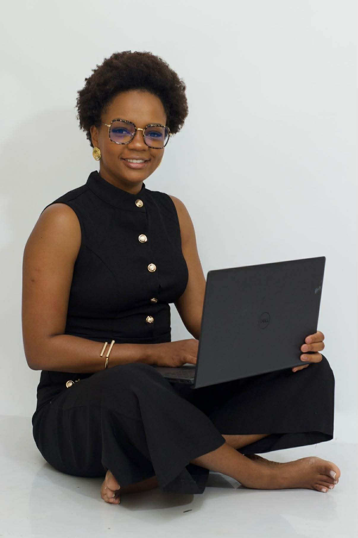 A woman sitting on the floor, holding a laptop, smiling, wearing glasses, gold earrings, a black sleeveless top with gold buttons, black pants, and a gold bracelet, against a plain white background.