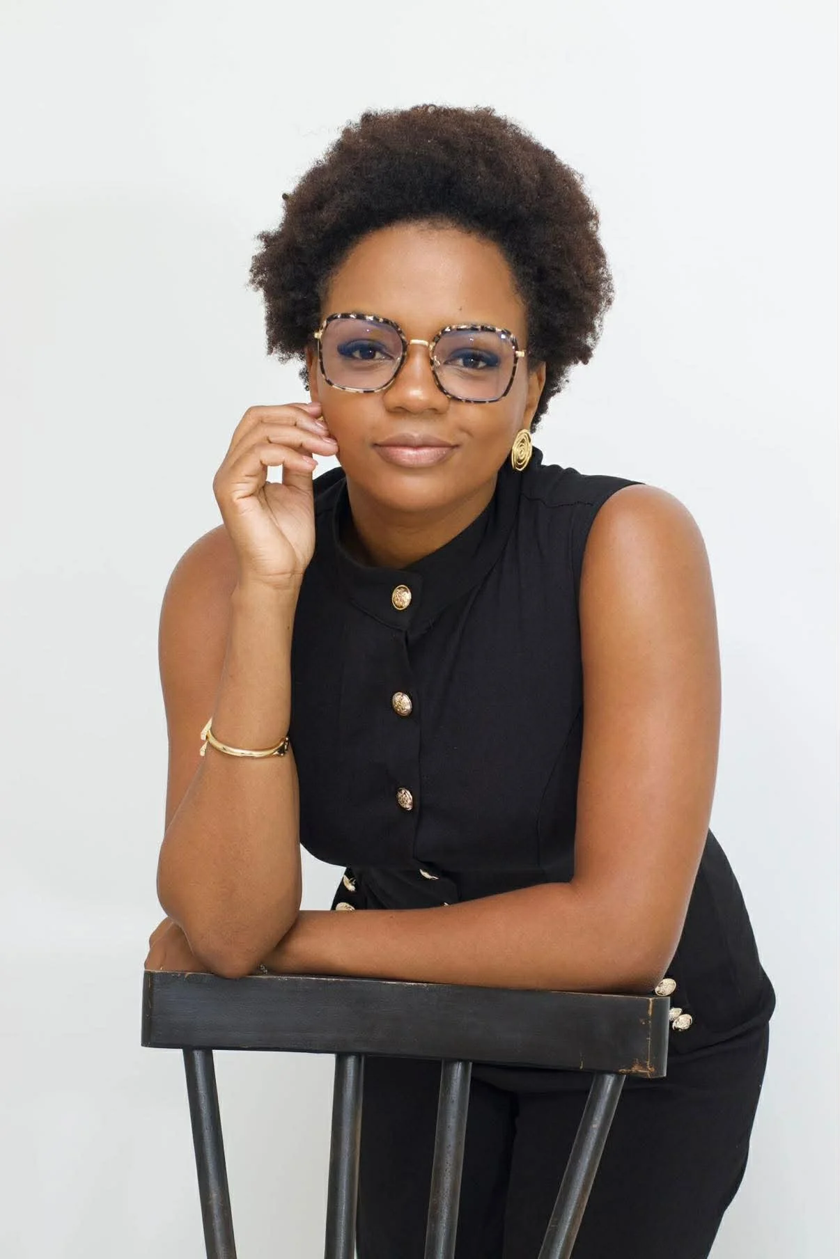 Portrait of a woman with curly hair and glasses, wearing a black sleeveless top, sitting on a chair without arms against a plain white background.