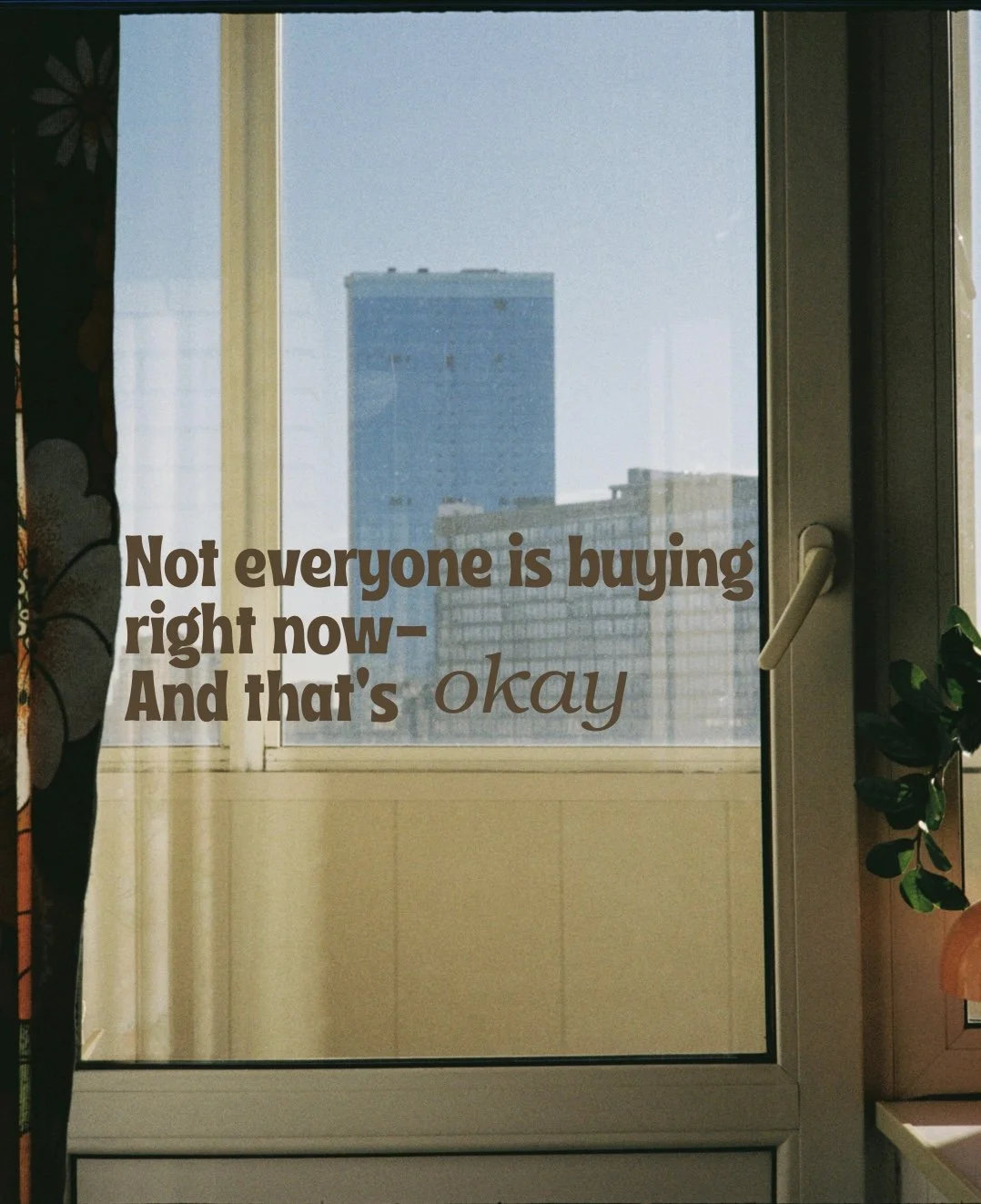 A view from inside a room looking out a glass door to a cityscape with tall buildings, with a motivational quote overlaid that says, "Not everyone is buying right now - And that's okay."