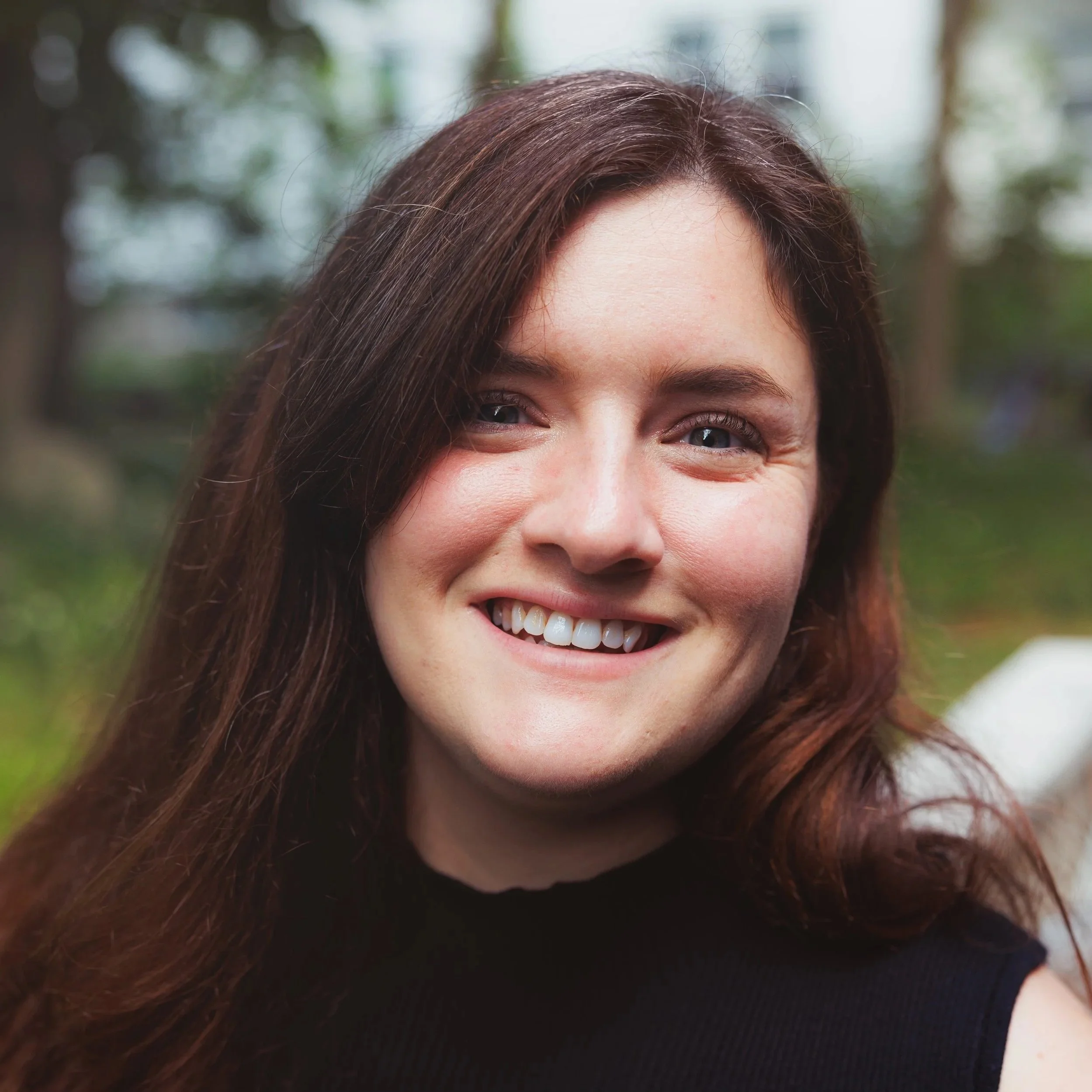 Partial view of a smiling woman with long brown hair and blue eyes outdoors, blurred trees in the background.