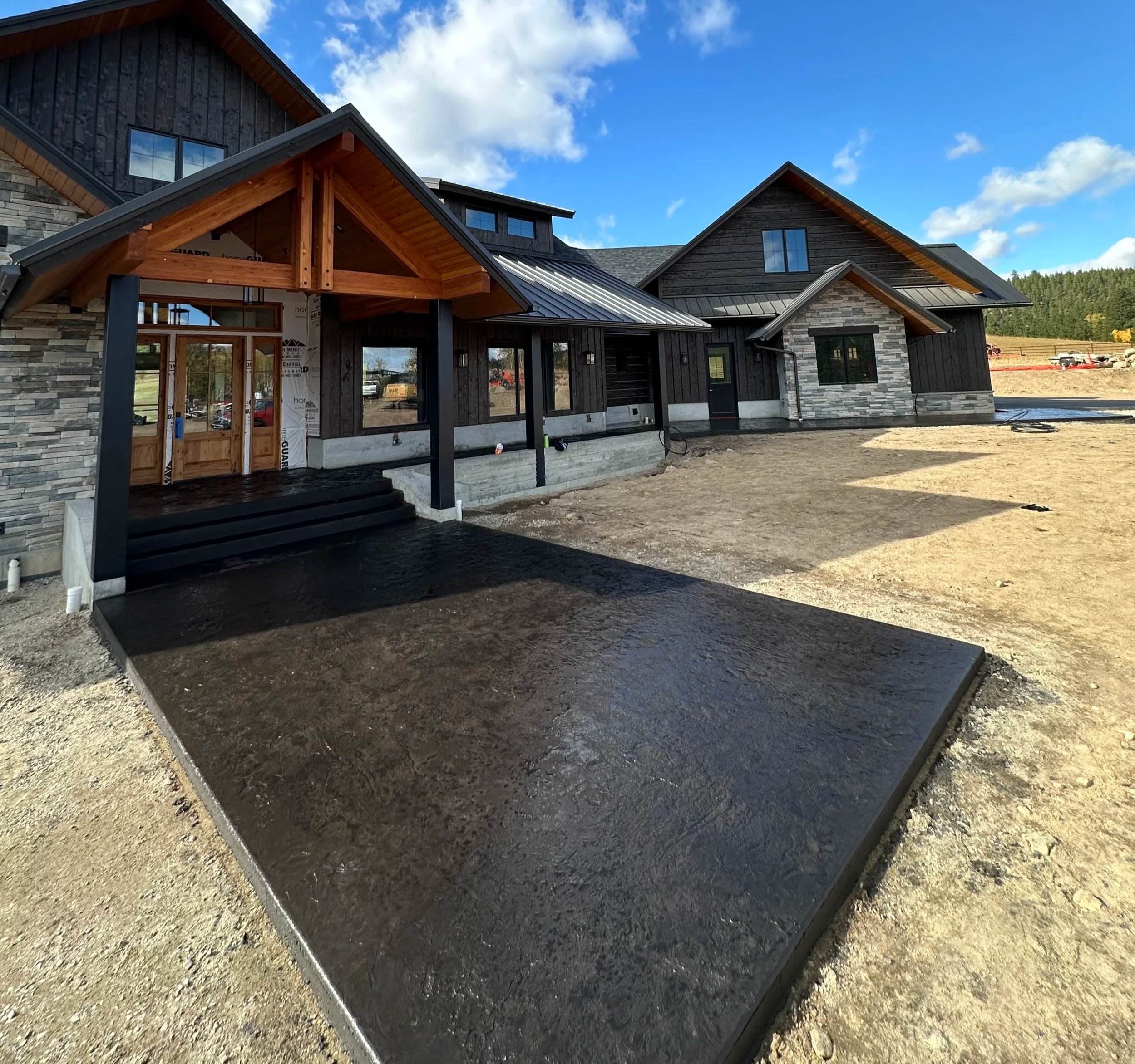 Newly constructed house with a combination of stone, wood, and metal exterior and a covered porch area, under a blue sky with clouds, with dirt and gravel around