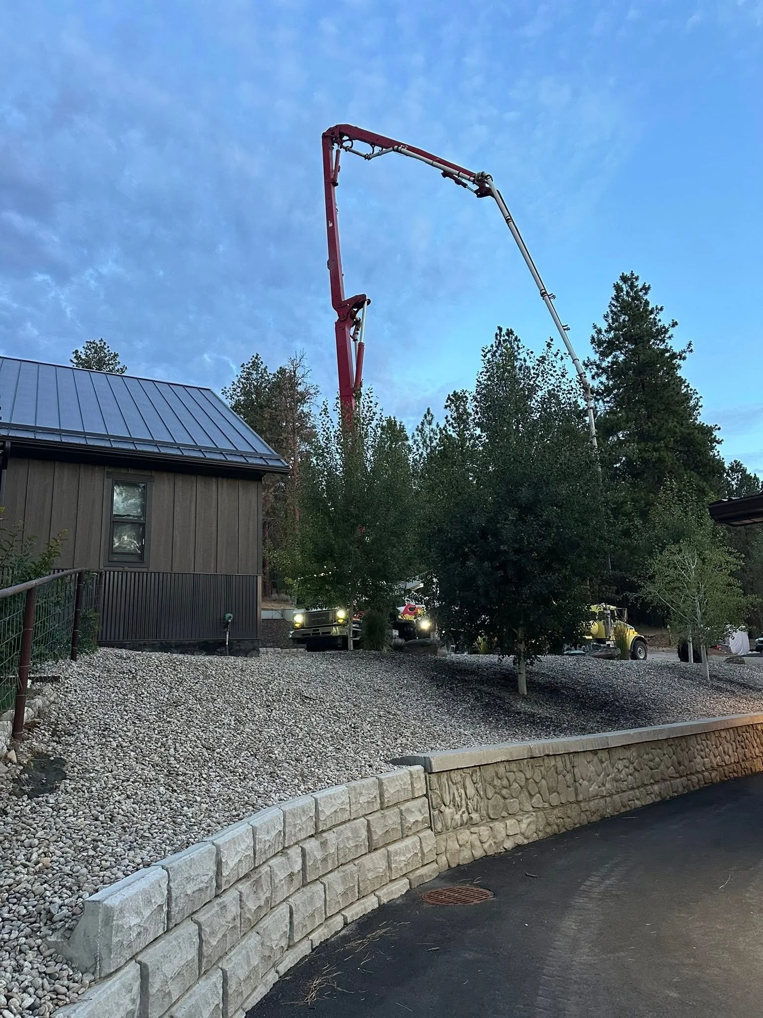 A construction scene at dusk showing a cement truck with a large red boom pump extending high into the sky, surrounded by trees and a building with a metal roof.