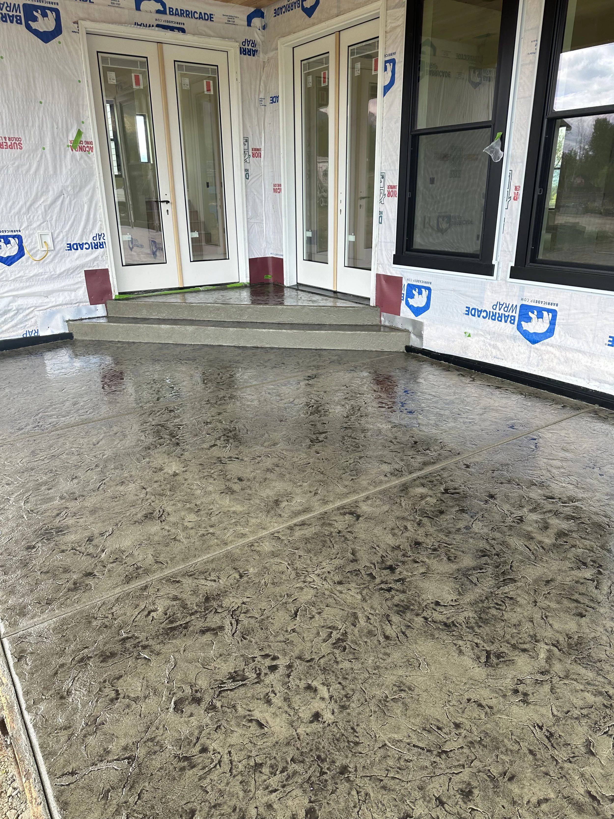 Porch area of a house under construction with concrete steps, unfinished walls covered with construction wrap, and large windows.